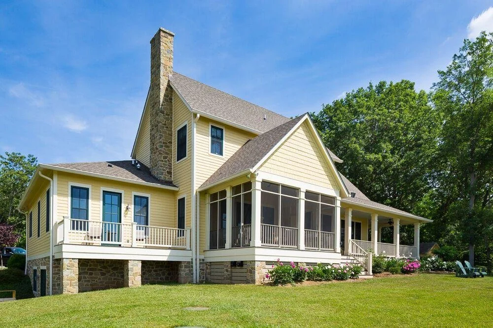 Yellow house with stone chimney, screened porch, and surrounded by green trees and lawn.