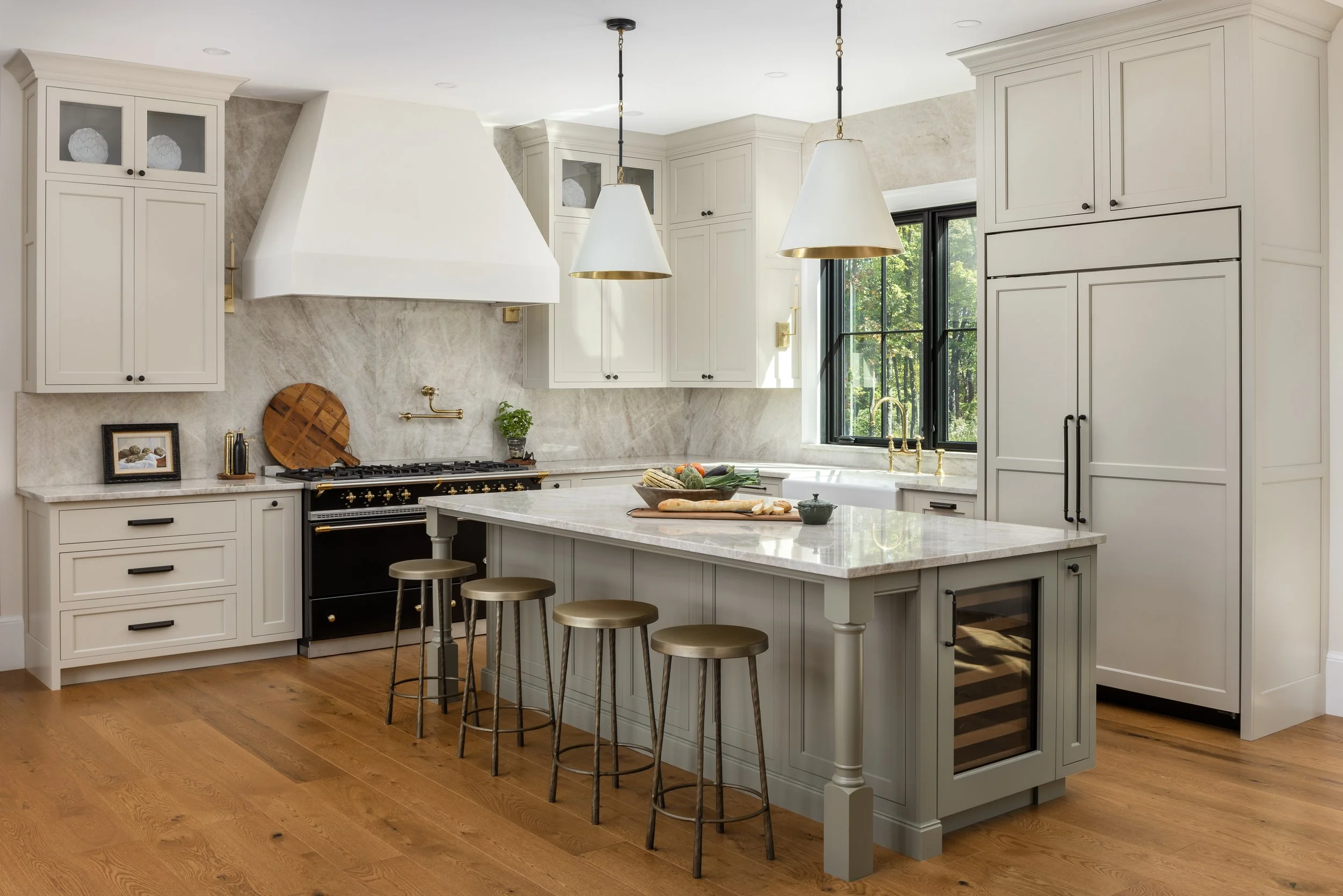 Modern kitchen with white cabinetry, marble countertops, wooden flooring, black and gold appliances, and three pendant lights over an island with barstools. A large window provides natural light, and there are decorative items and kitchen tools on th