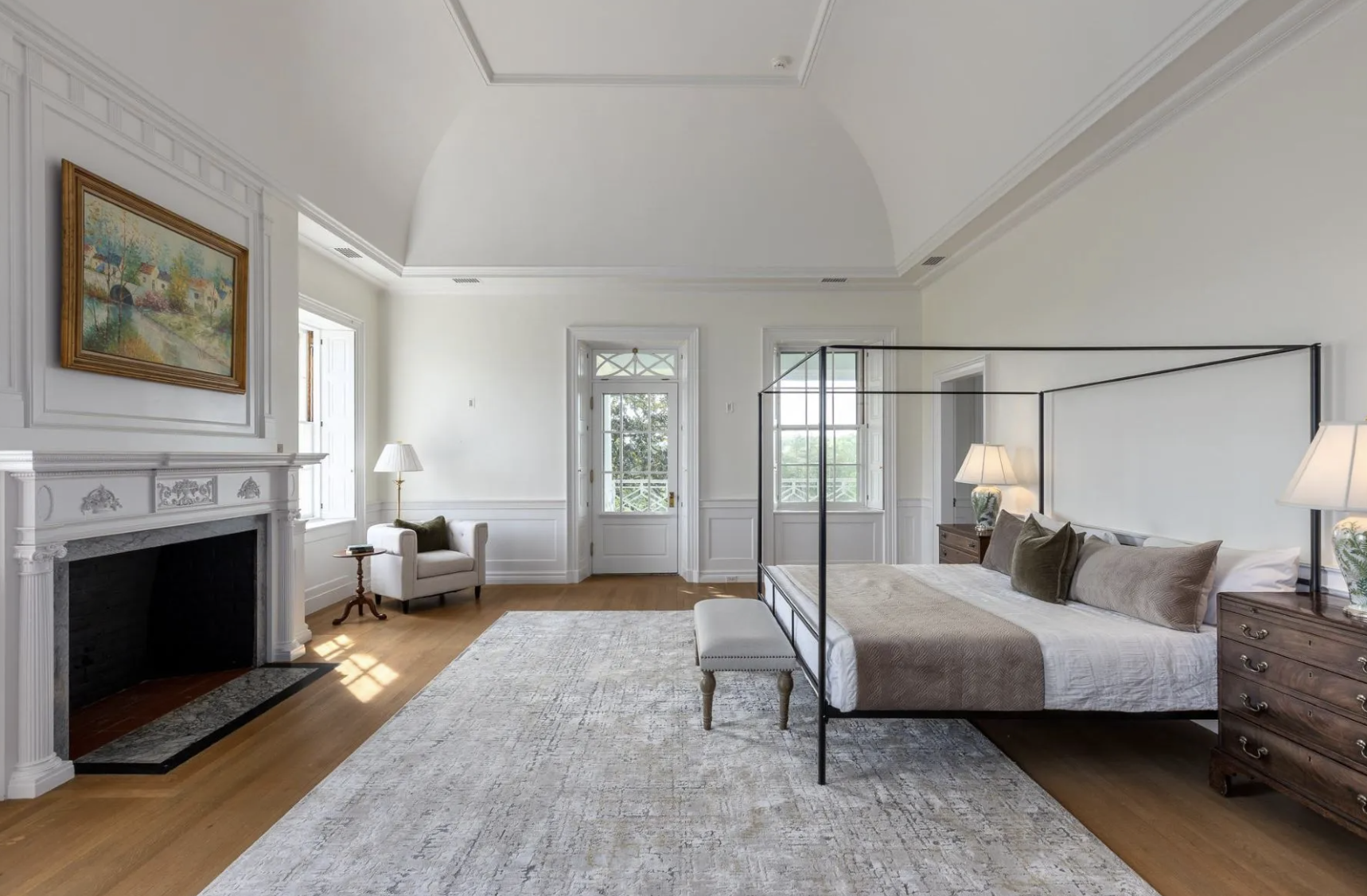 Spacious bedroom with a white fireplace, a large bed with brown and beige pillows, nightstands with lamps, a rug, a small bench at the foot of the bed, and large windows with a view of trees.