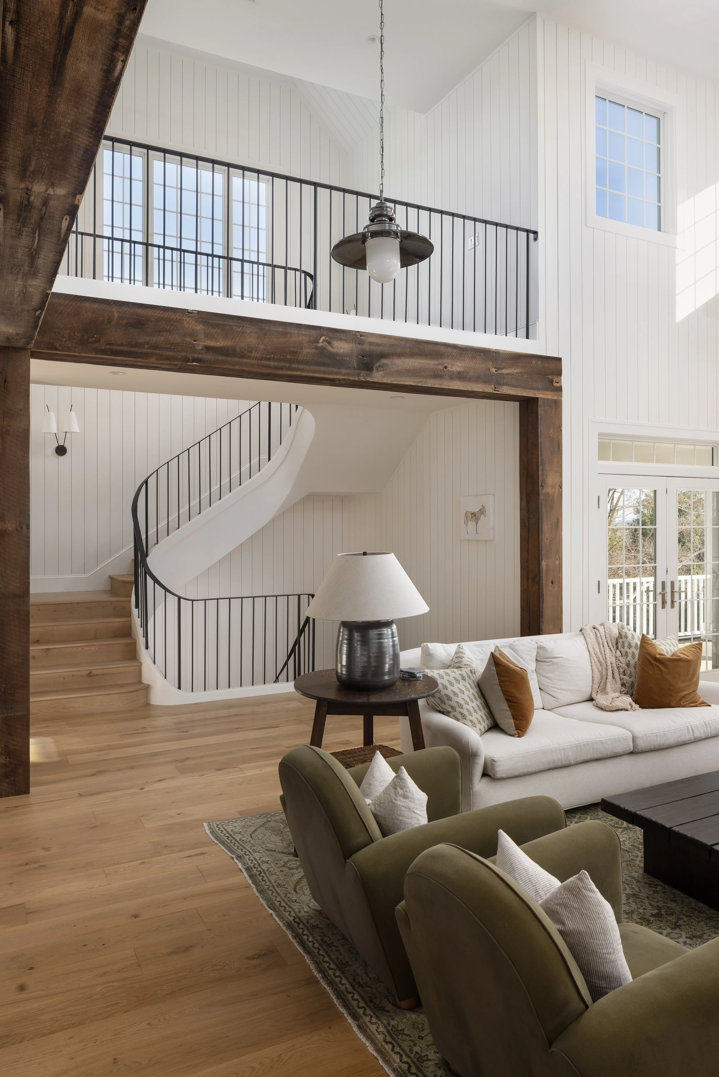Living room with white coiled sofa, green armchairs, wooden coffee table, floor lamp, and staircase leading to upper floor. Large windows and sliding glass doors let in natural light.