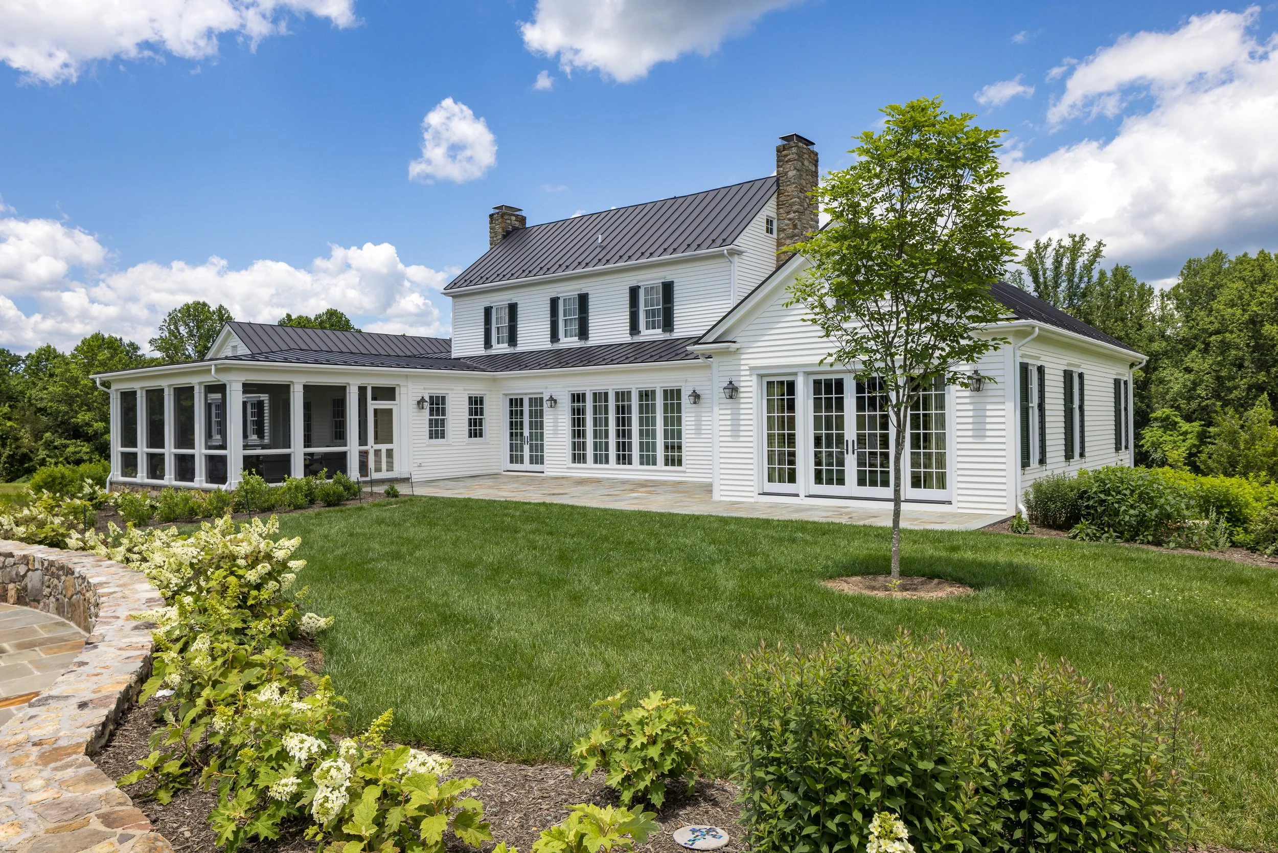 A large white house with black shutters, a stone chimney, multiple glass doors and windows, a screened porch, and a well-maintained lawn with trees and shrubs under a partly cloudy blue sky.