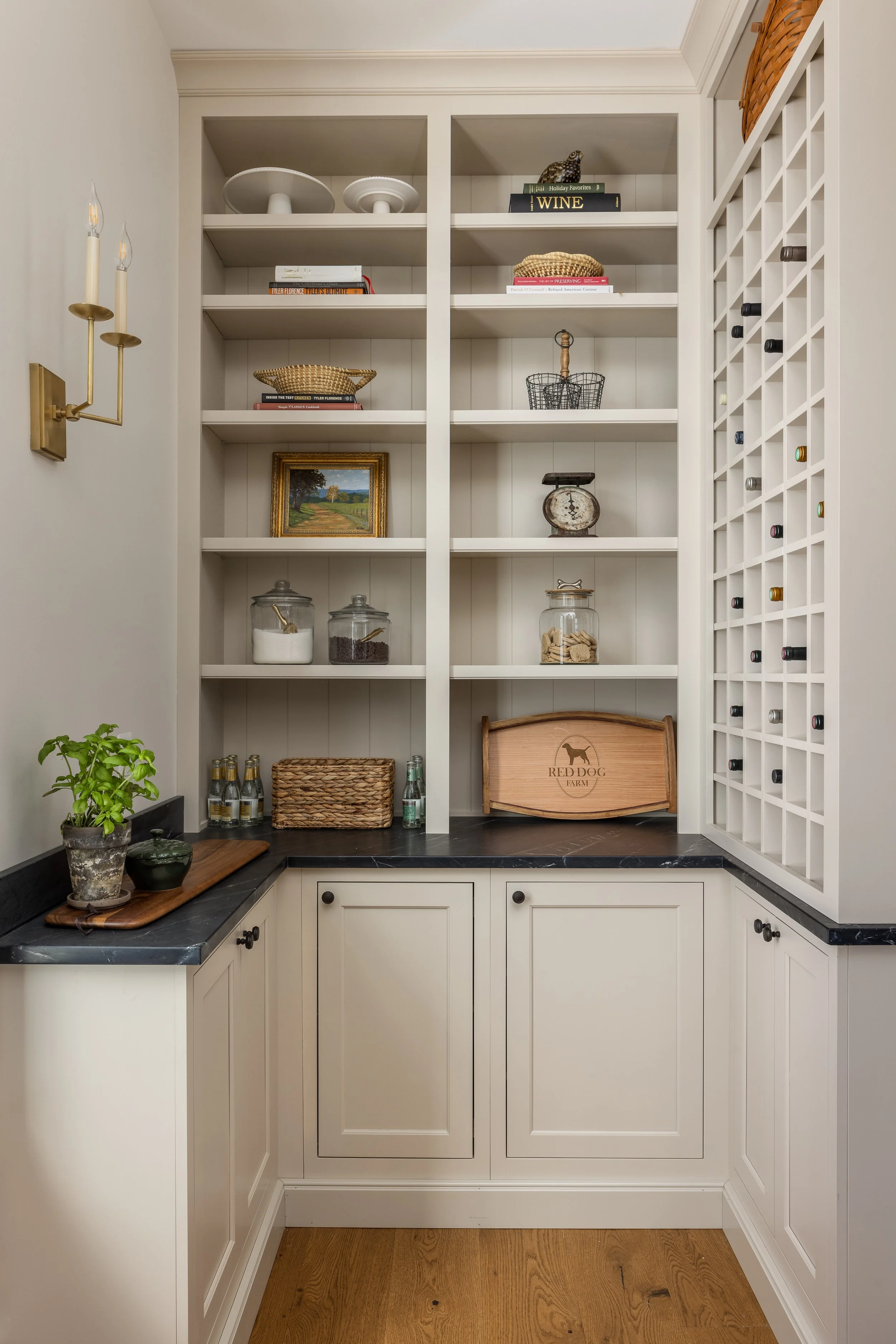 A built-in white kitchen storage cabinet with open shelves holding books, baskets, and decorative items, and a wine rack on the side, with a black countertop and a small plant on the corner.