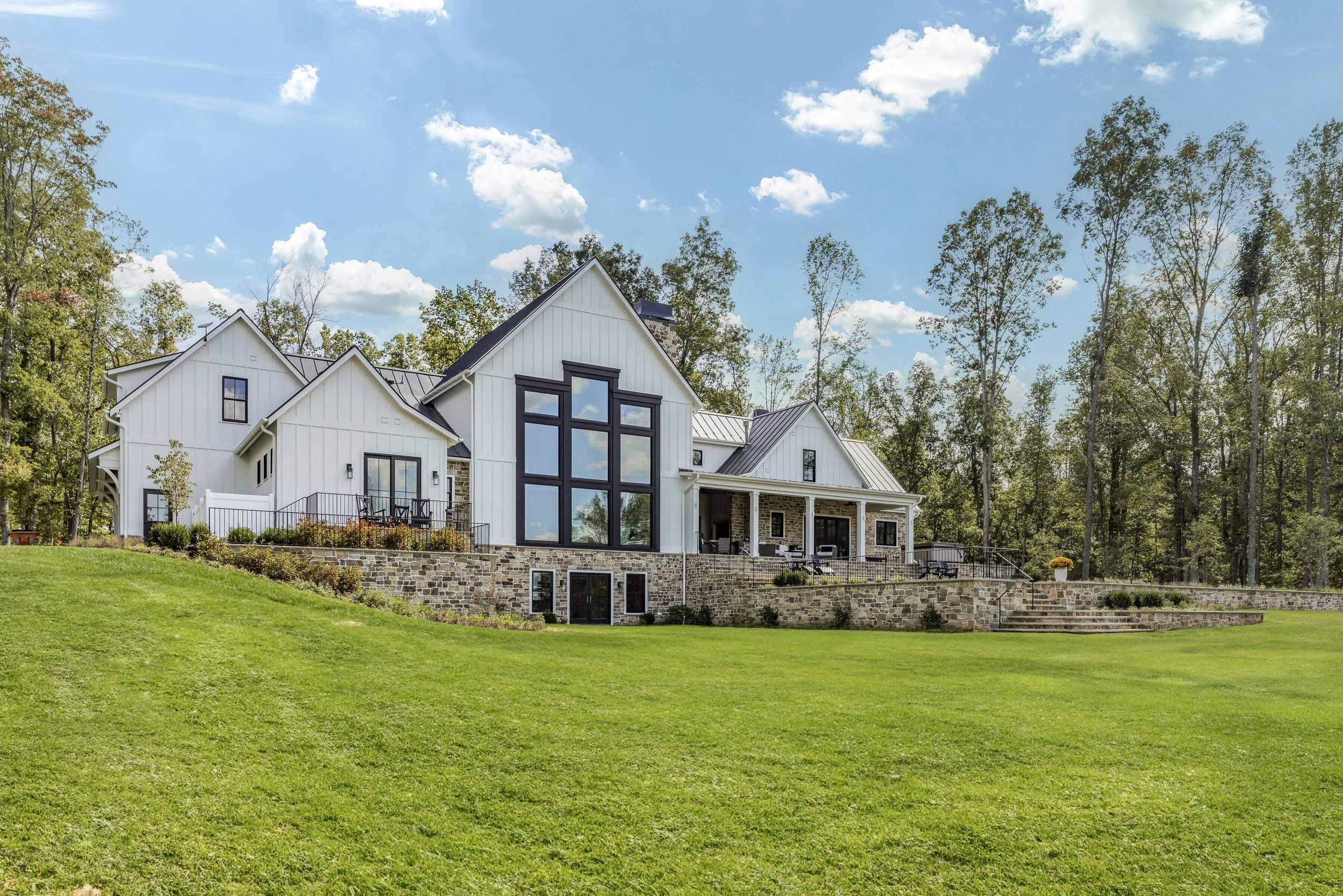 Large modern house with white siding and black window frames, built on a stone foundation, surrounded by green lawn and trees, under a bright blue sky with clouds.