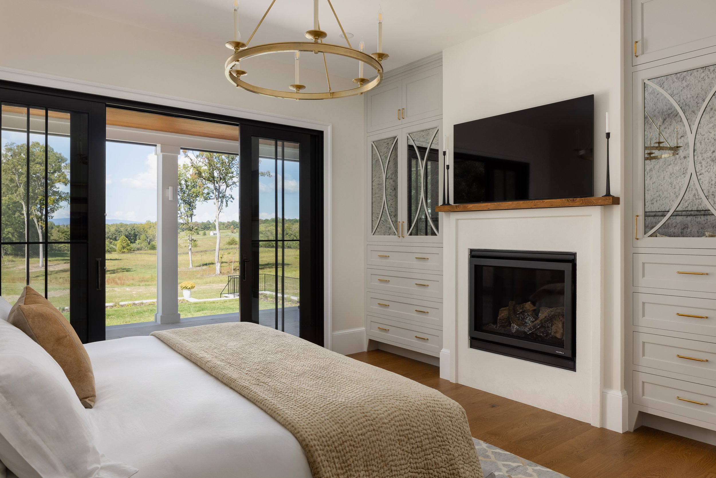 Living room with white walls, a fireplace, a wall-mounted TV, built-in cabinets with glass doors, a large glass sliding door showing a green outdoor landscape with trees, and a gold circular chandelier.