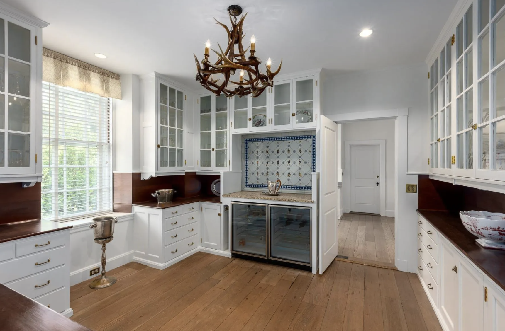 A kitchen with white cabinets, glass-front upper cabinets, wooden countertops, a window with blinds, and a chandelier made of antlers.