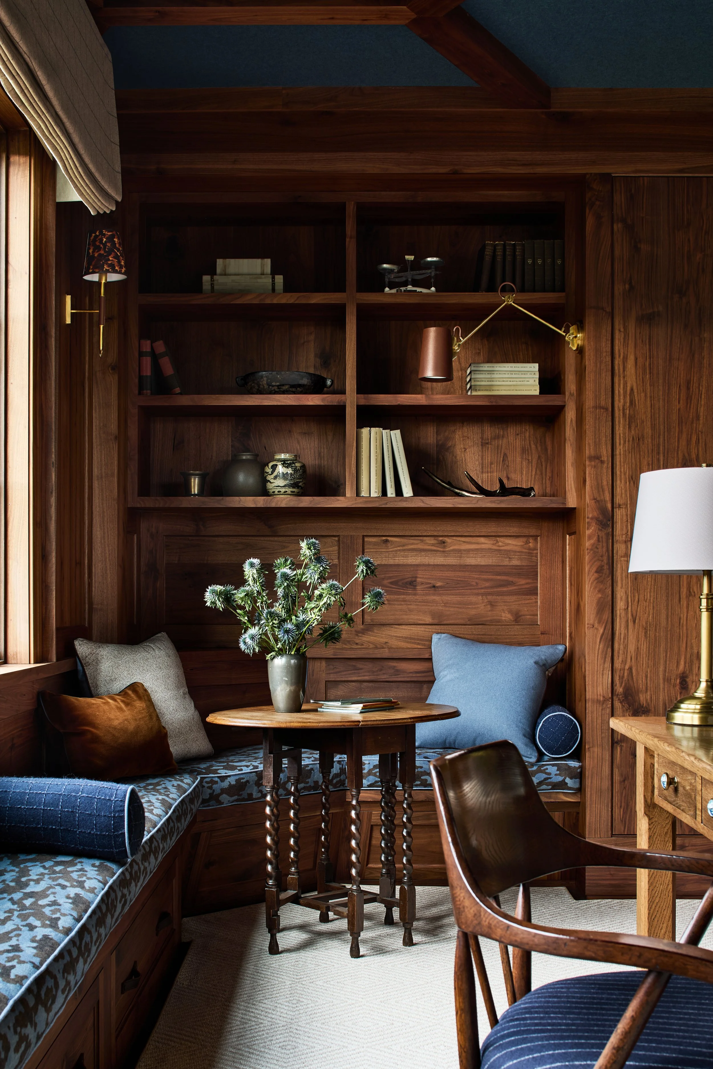 A cozy corner with built-in wooden bench seating, decorative pillows, a small round wooden table with a flower vase, and a bookshelf with decorative items, books, and a wall-mounted lamp