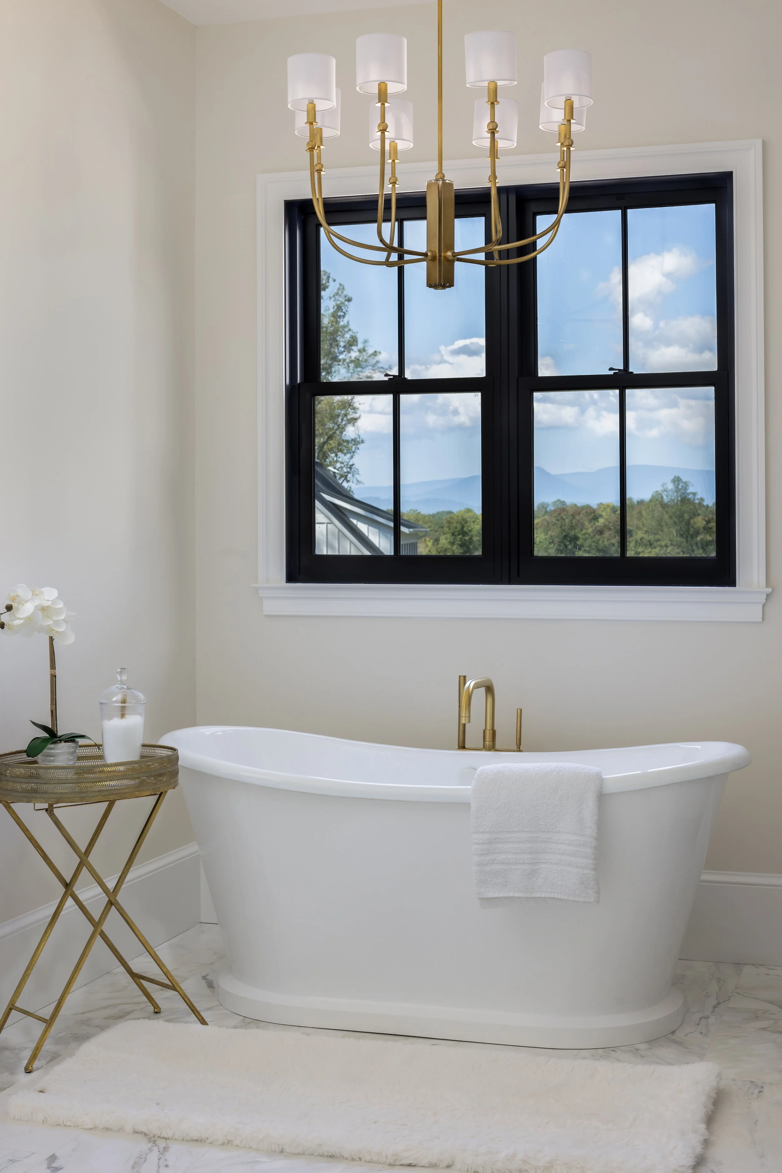 A modern bathroom with a white bathtub, gold fixtures, a large black-framed window showing a mountain view, a gold chandelier with white lampshades, a side table with a plant and decorative items, and a white shaggy rug.