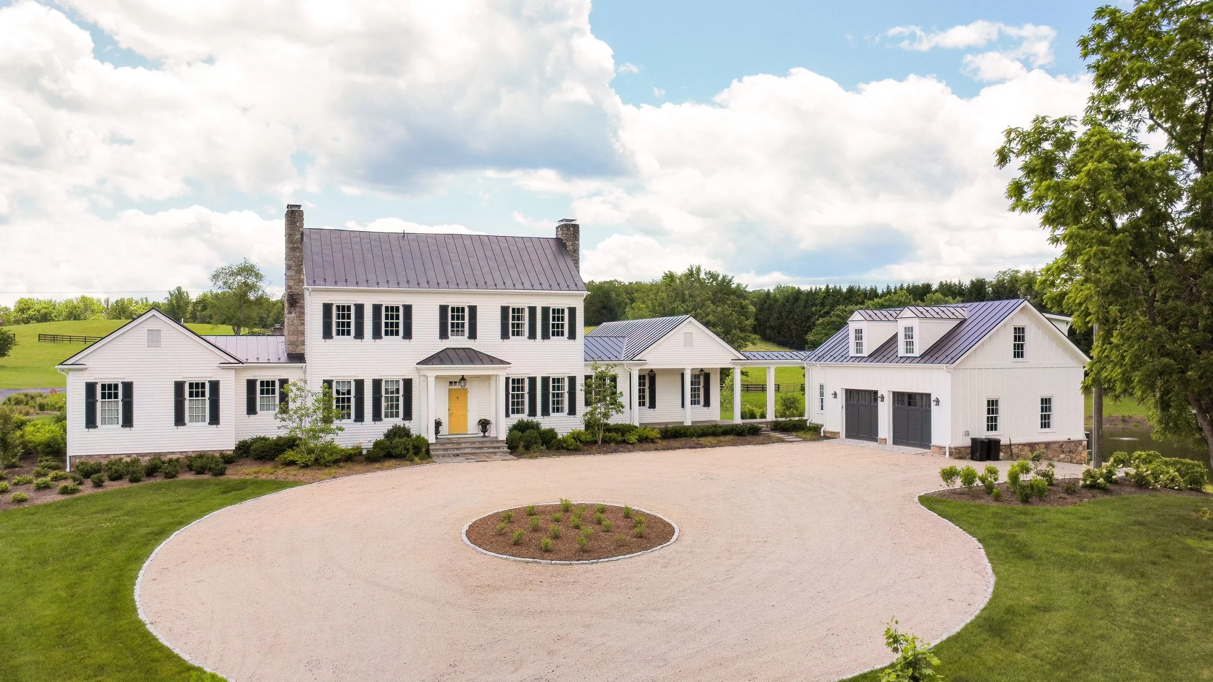 A large white country house with black shutters, a yellow front door, and a metal roof. A detached garage with black doors is nearby, and a circular driveway with a garden bed in the center surrounds the front yard. Trees and green hills are in the b