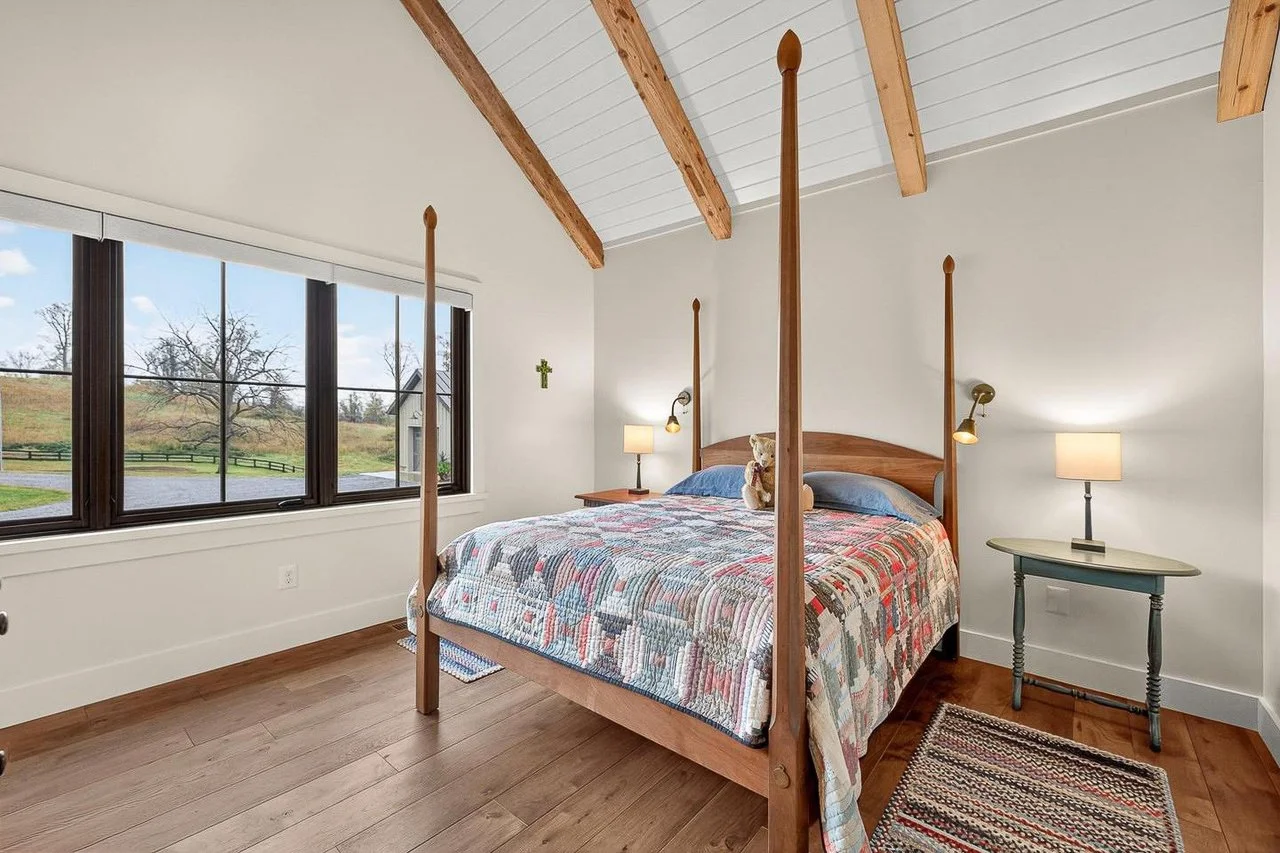 A cozy bedroom with a wooden four-poster bed, patchwork quilt, stuffed bear, two bedside lamps, small table, large window with a view of trees and grass outside, and a colorful striped rug.