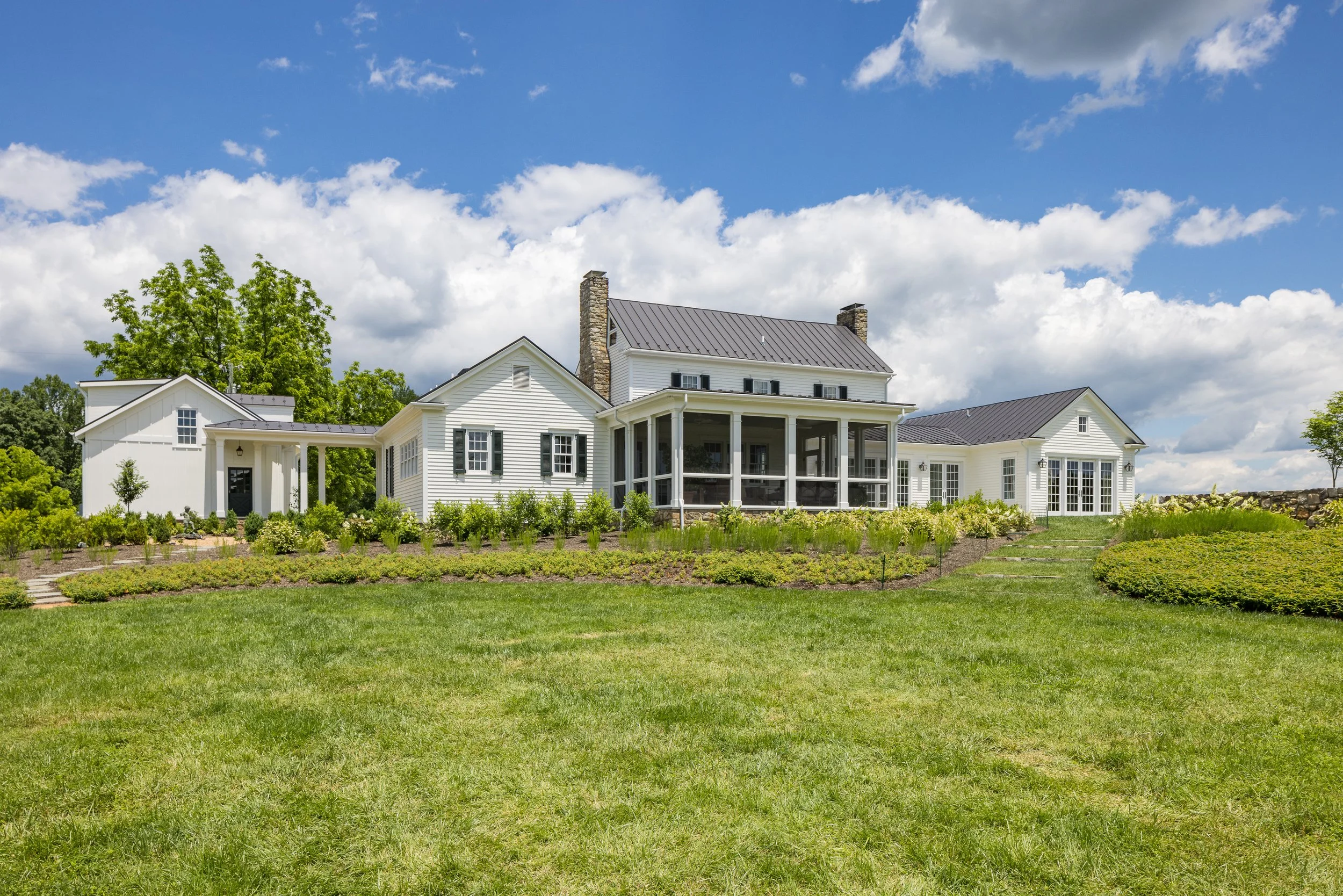 A large white house with a dark metal roof and a screened porch, set on a lush green lawn with landscaped garden beds, under a partly cloudy blue sky.
