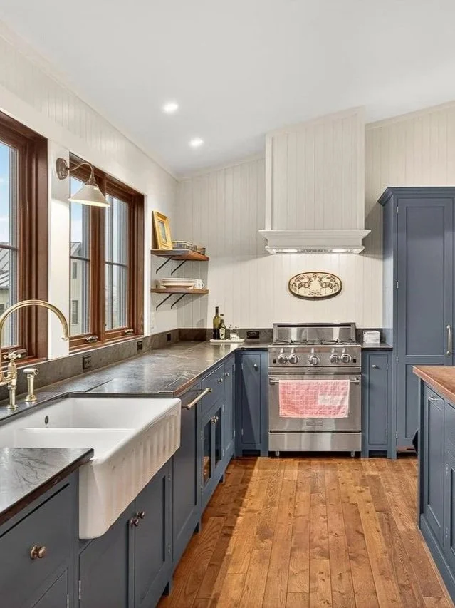 Kitchen with blue cabinets, a farmhouse sink, a stainless steel stove, wooden floors, and large windows.