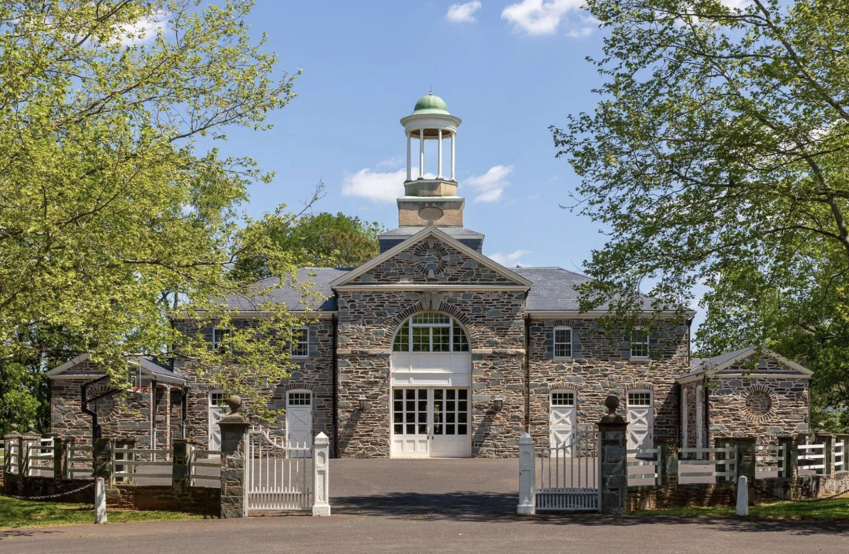 Stone building with a white gate, trees, and a clock tower with a dome, under a blue sky.