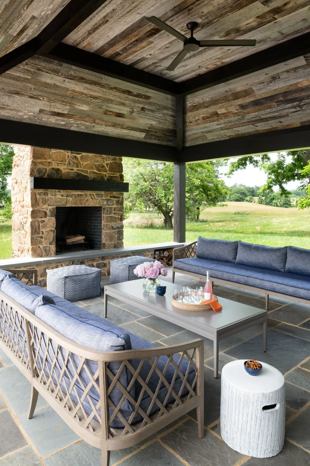 Covered outdoor patio with stone fireplace, wooden ceiling, ceiling fan, blue cushioned sofas, patterned ottomans, white coffee table with pink flowers, wine and glasses, and a small stone side table with a bowl of snacks, overlooking a green field a