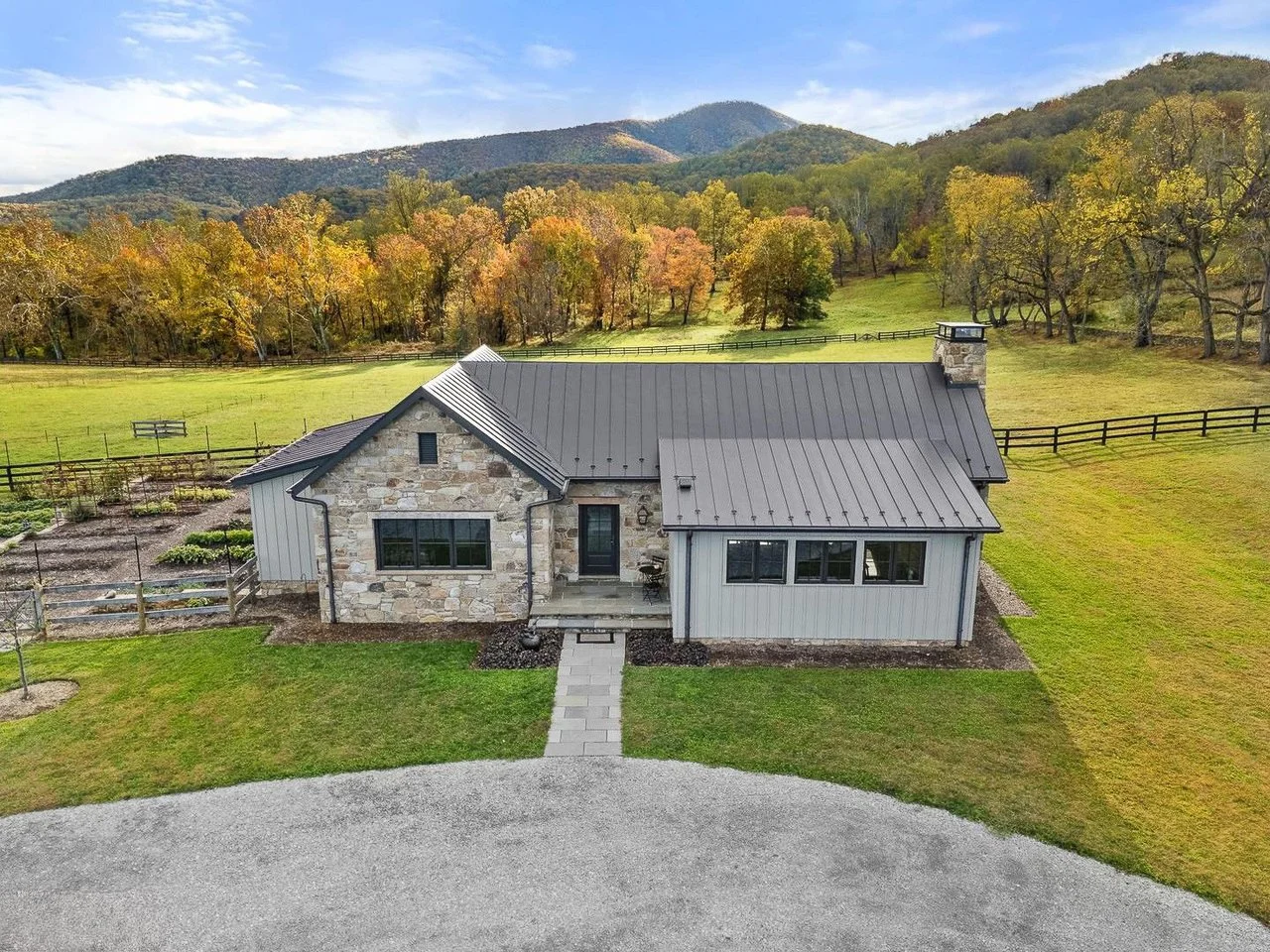 A house with a stone and metal exterior, surrounded by a green lawn and fenced gardens, with trees and mountains in the background under a partly cloudy sky.