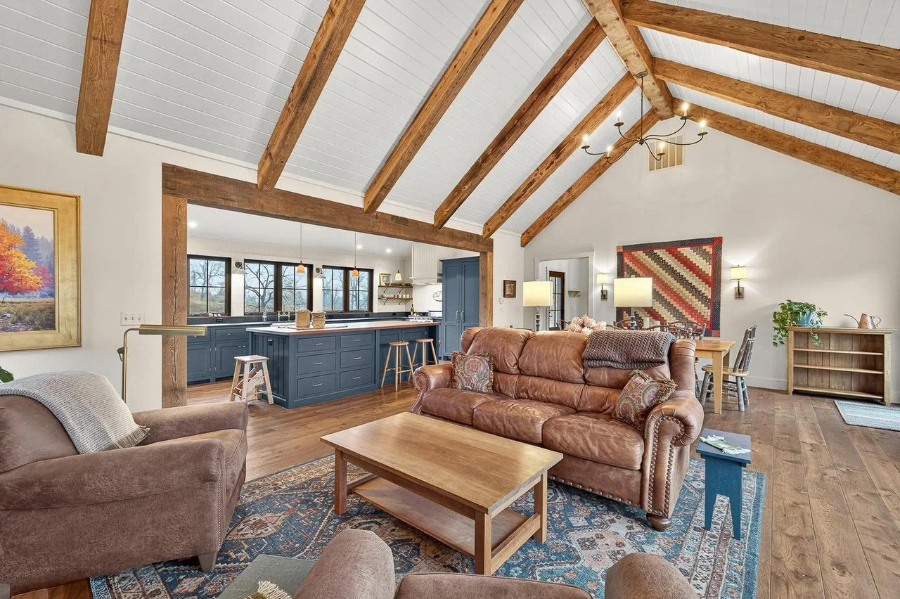 A spacious living room with vaulted ceiling, exposed wooden beams, and hardwood floors. It features a brown leather sofa, a matching armchair, and a wooden coffee table on an ornate rug. The room extends into a kitchen area with blue cabinetry and a 