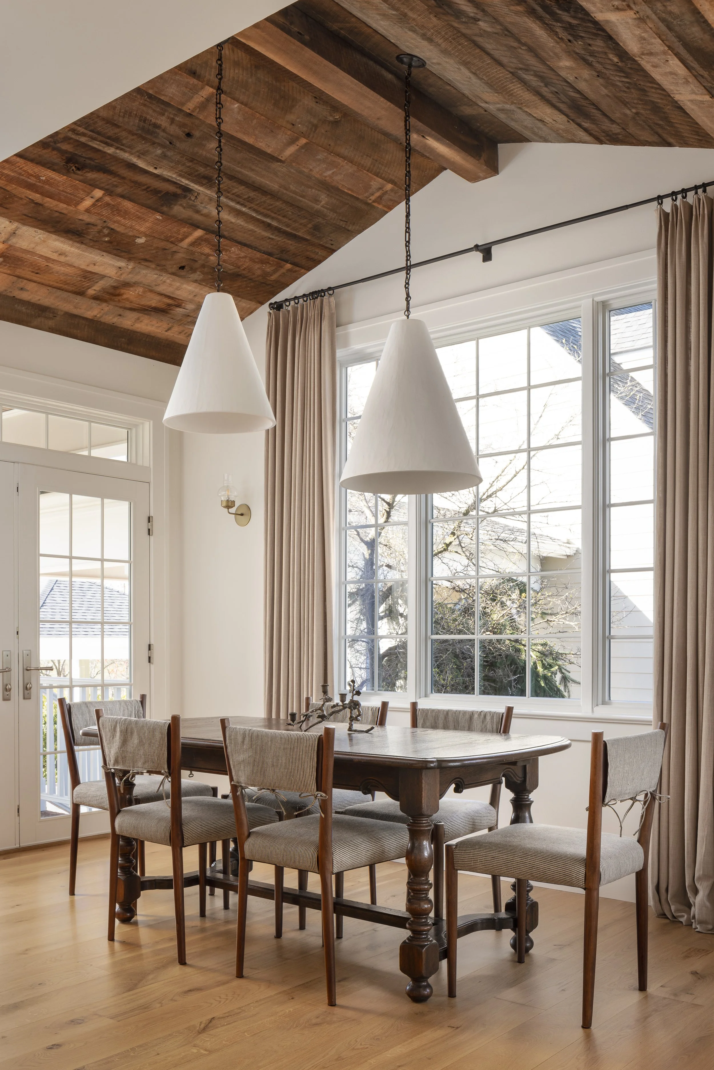 A dining room with a wooden table surrounded by six upholstered chairs, large windows with beige curtains, a wooden ceiling with hanging white pendant lights, and a glass door leading to an outdoor space.