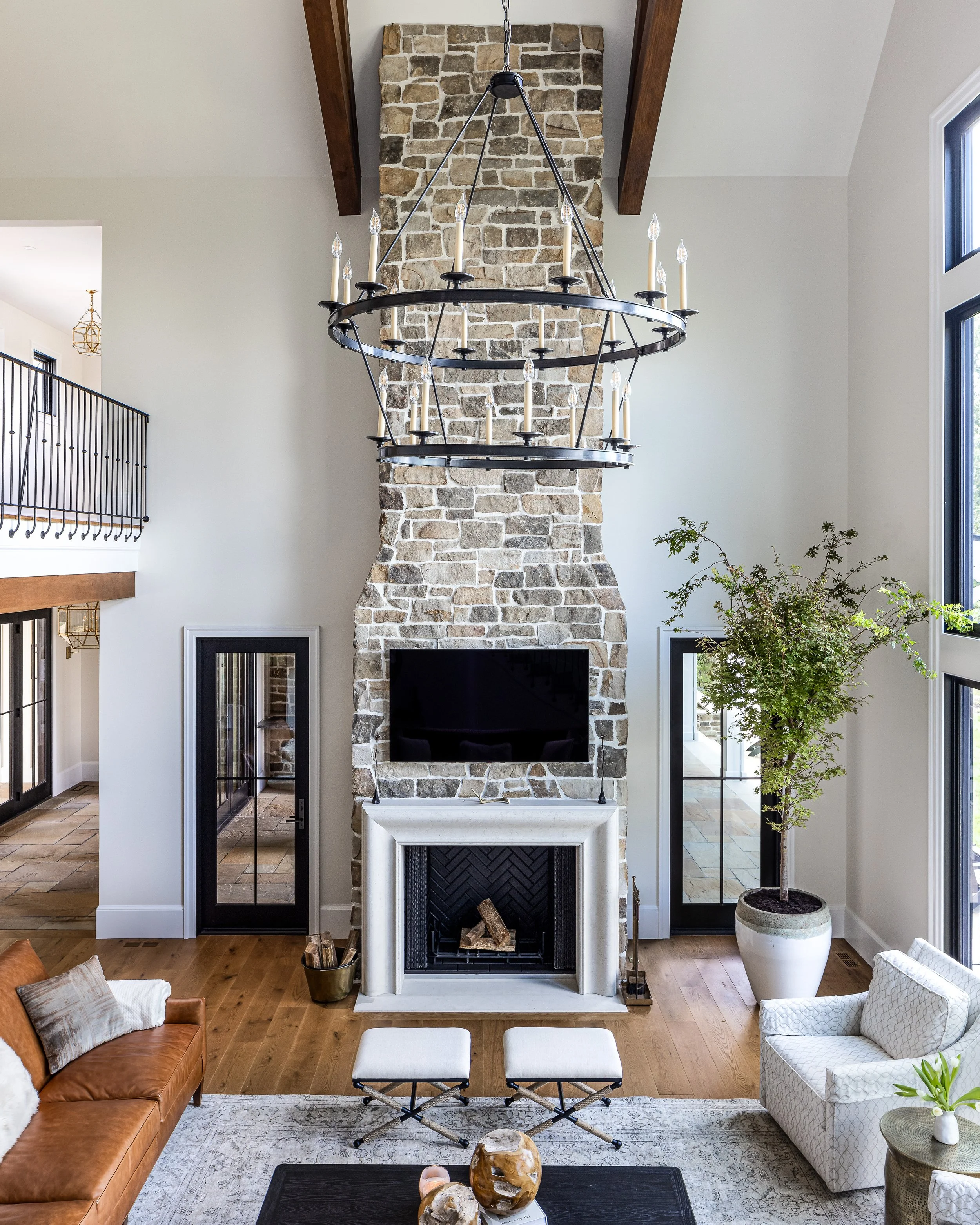 Living room with a tall stone fireplace, mounted television, large window, potted tree, leather sofa, armchair, and decorative vases.