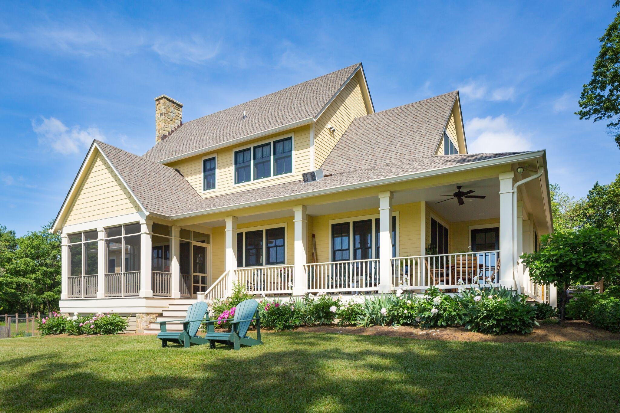 Yellow multi-story house with a porch, surrounded by green lawn and bushes, under a blue sky with some clouds.