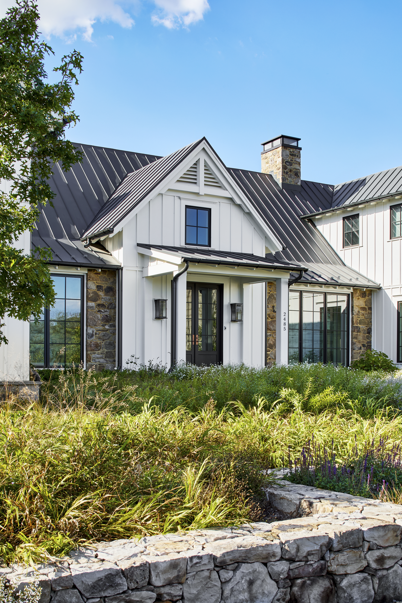 A modern white house with black trim, stone accents, and large windows, surrounded by a garden with green plants and a stone border, under a blue sky with a few clouds.