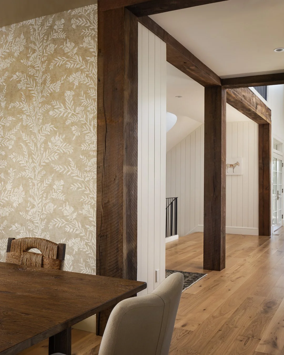 Interior of a home featuring a wooden dining table, upholstered chairs, a wallpapered wall with a leafy pattern, exposed wooden beams, vinyl plank flooring, and a hallway with a framed animal painting.