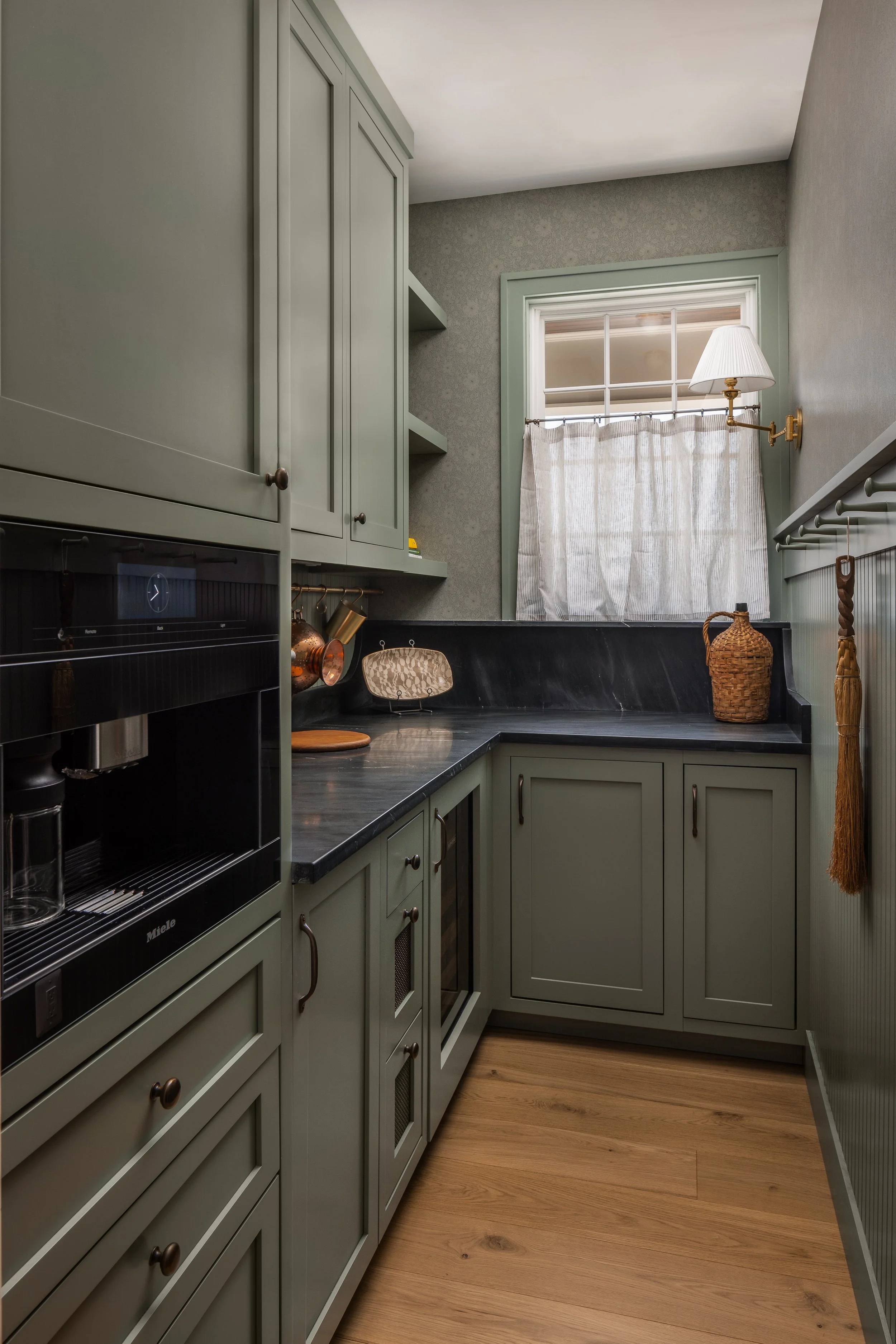 A small kitchen with green cabinets, black countertop, a window with a curtain, and wooden flooring.