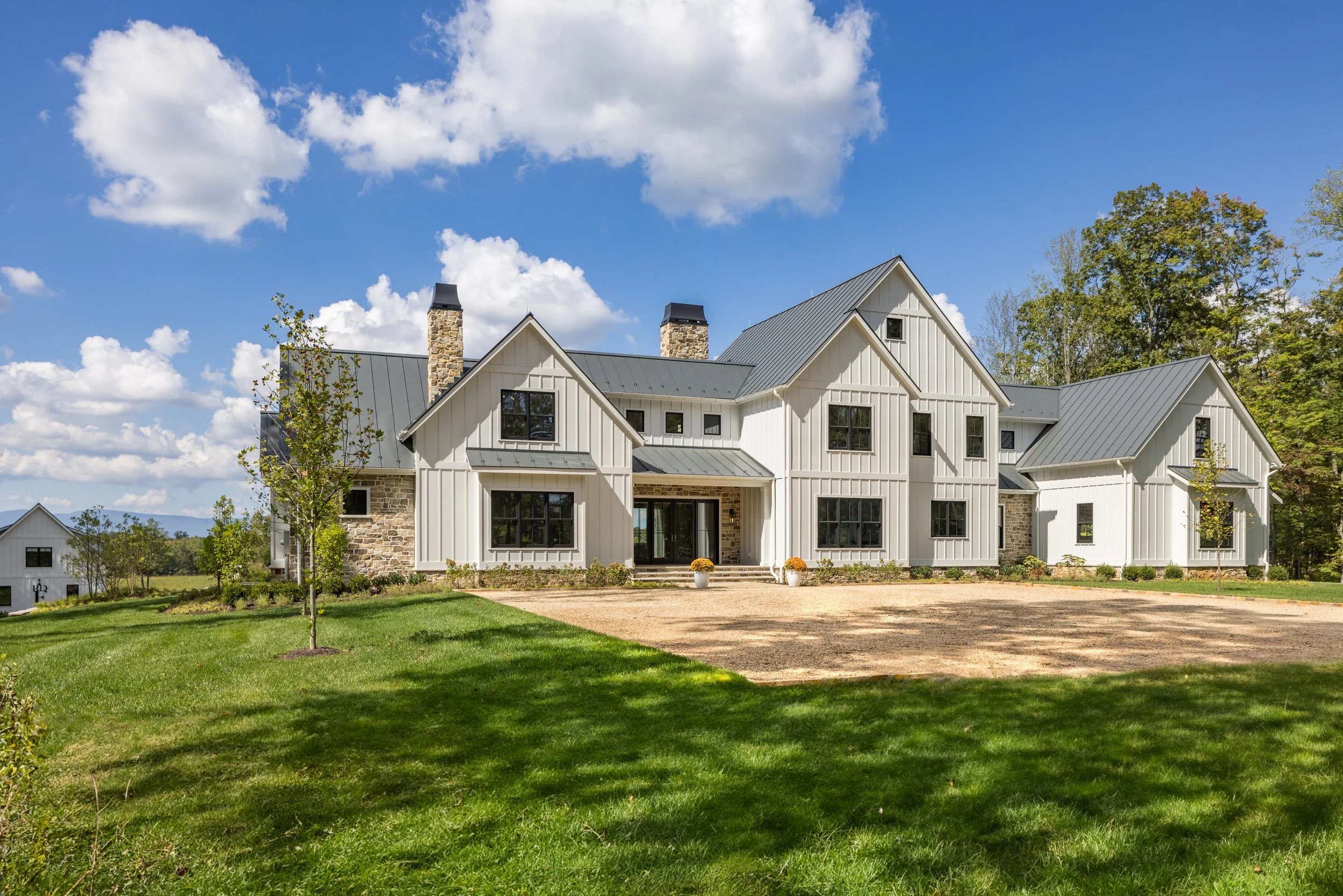 A large white house with black window frames and a metal roof, surrounded by green grass and trees, under a partly cloudy sky.