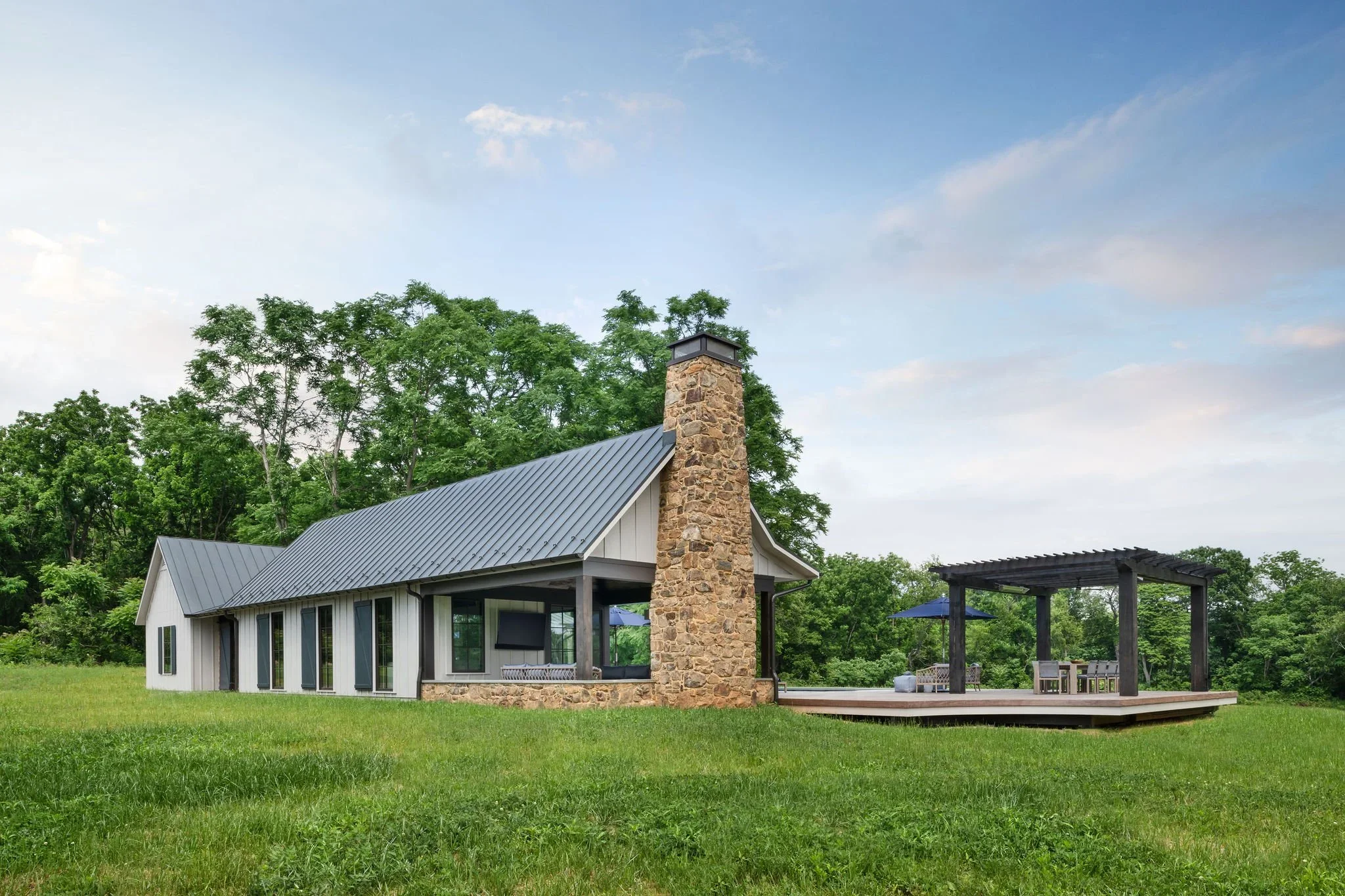 Modern house with metal roof and stone chimney on grassy lawn with trees in the background.