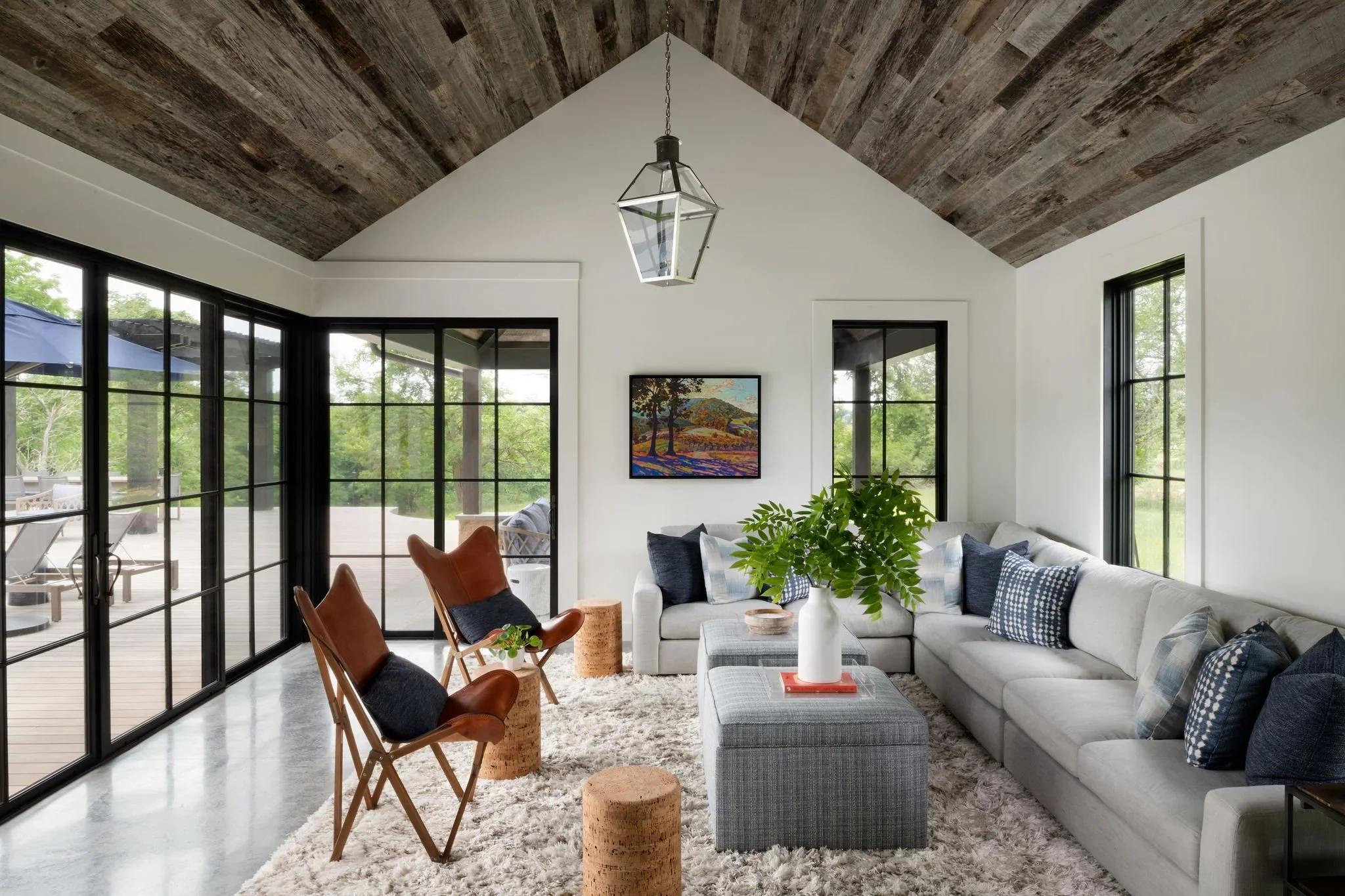 Bright living room with a vaulted wooden ceiling, large black-framed windows, a sectional sofa with blue and white throw pillows, a rectangular coffee table with a white vase and greenery, two leather and wood chairs, cork side tables, a textured whi