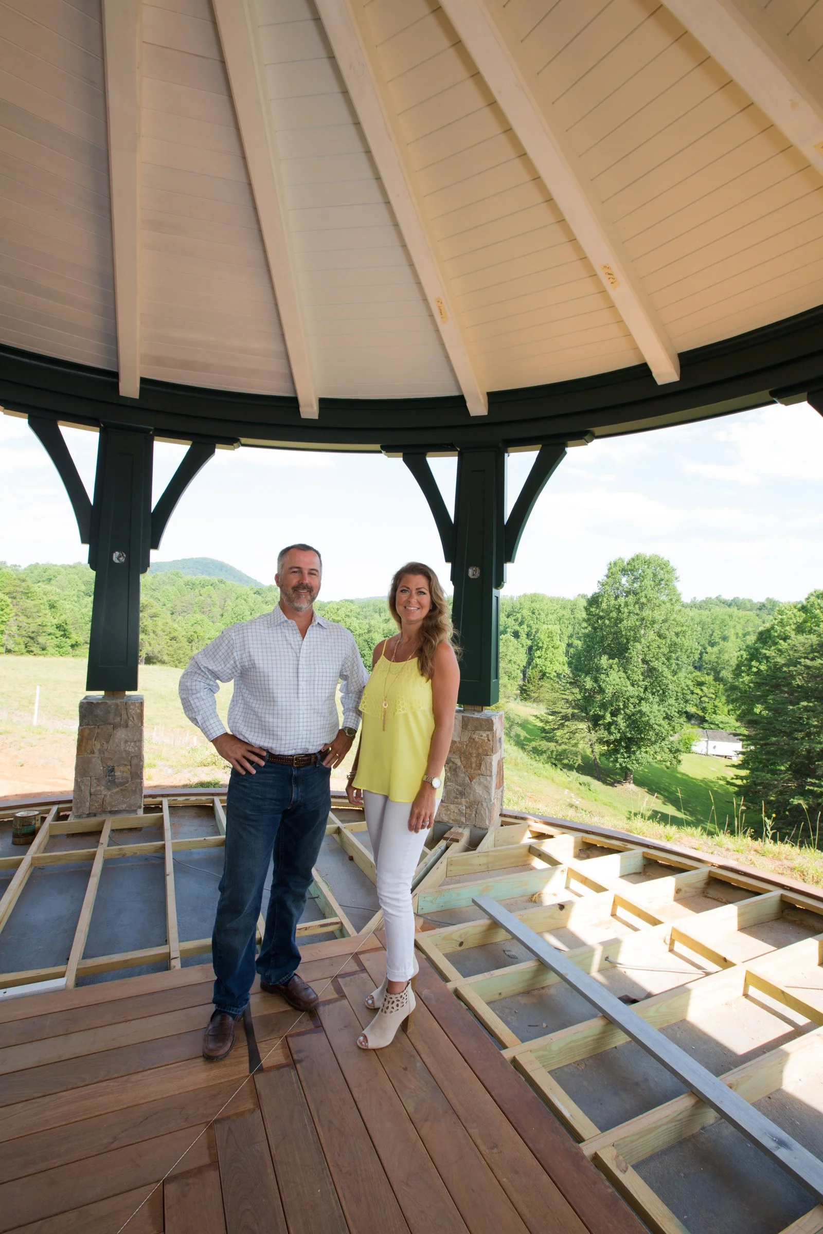 A man and a woman standing outdoors on a deck under a partially built gazebo with a scenic green landscape in the background.