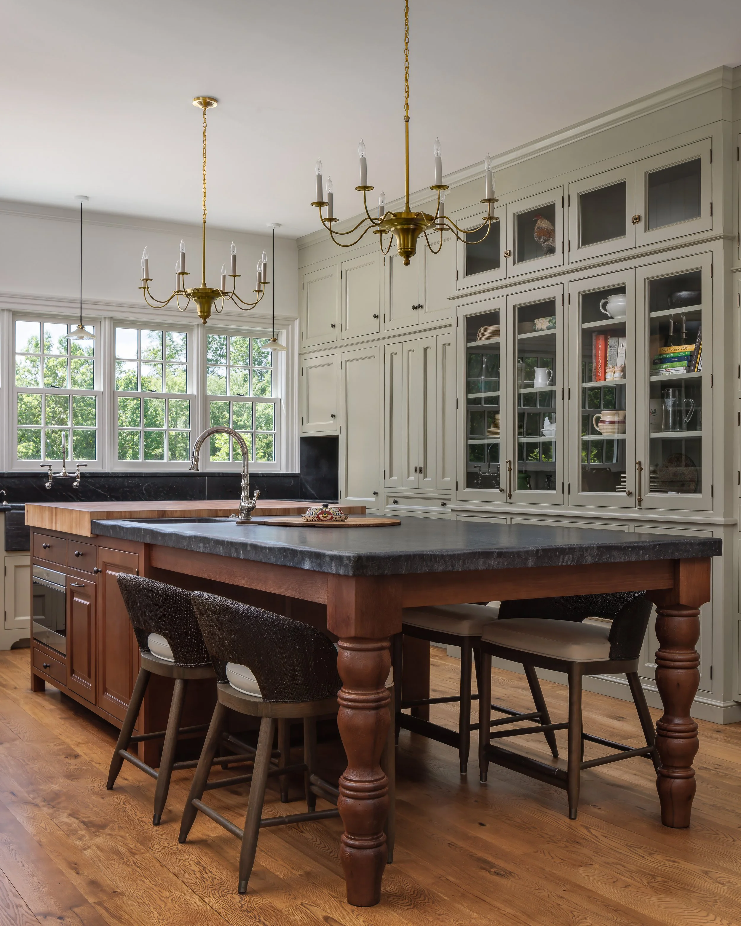 Kitchen with a large wooden island, black countertop, white cabinets with glass doors, a window with greenery outside, and two hanging chandeliers.