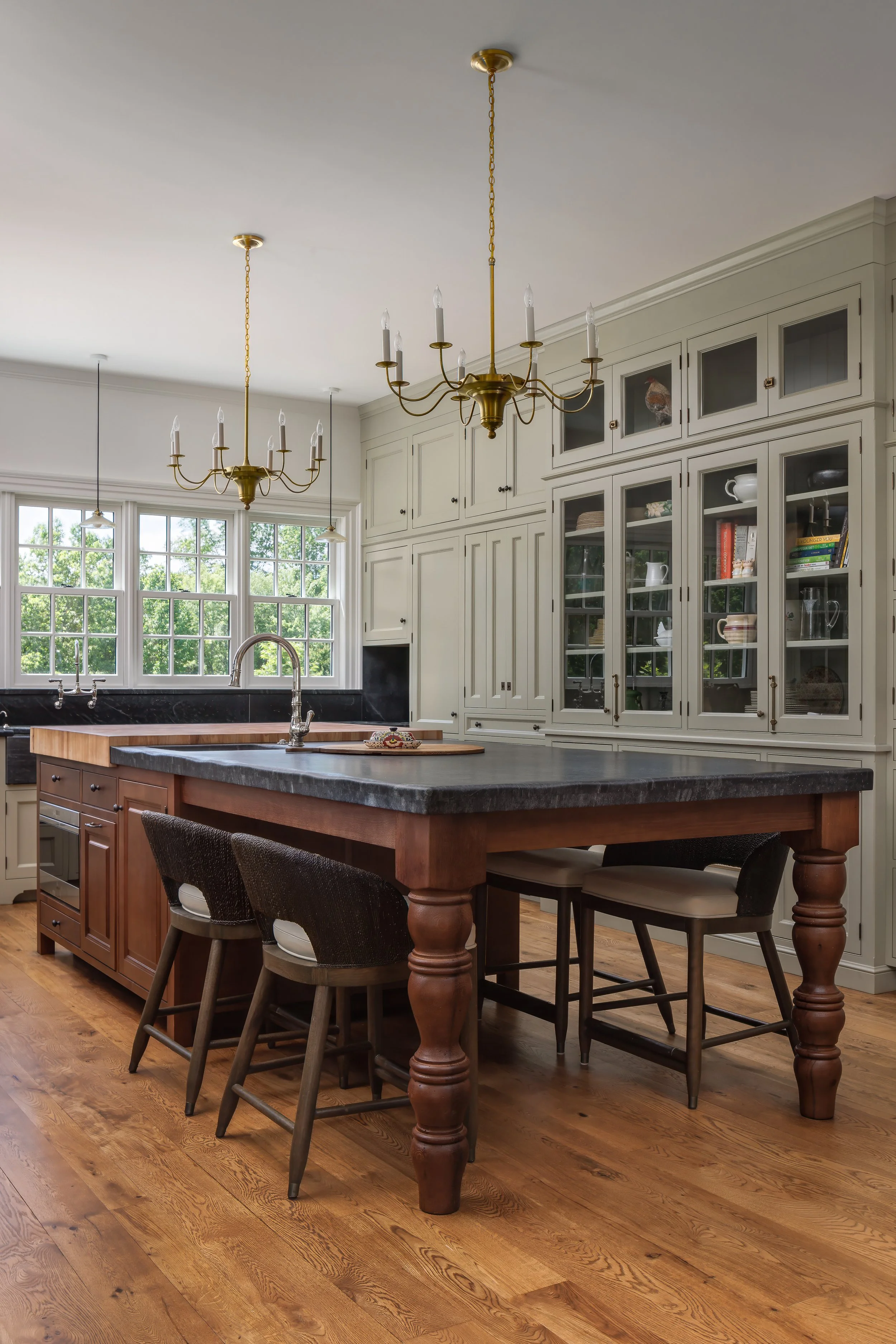 A spacious kitchen with a wooden island and chairs, white cabinets with glass doors, large windows, and two chandeliers hanging from the ceiling.
