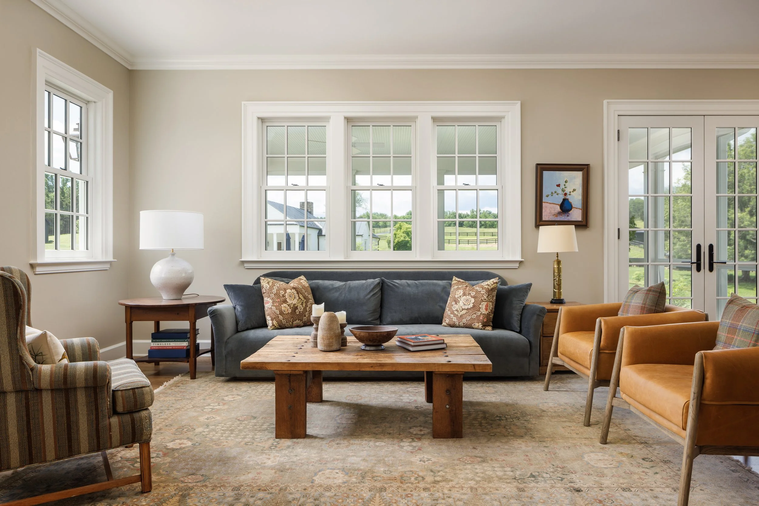 Living room with beige walls, multiple windows, a gray sofa with patterned pillows, two tan armchairs with plaid pillows, a rustic wooden coffee table, side tables with lamps, and a framed artwork on the wall, overlooking a green outdoor landscape.
