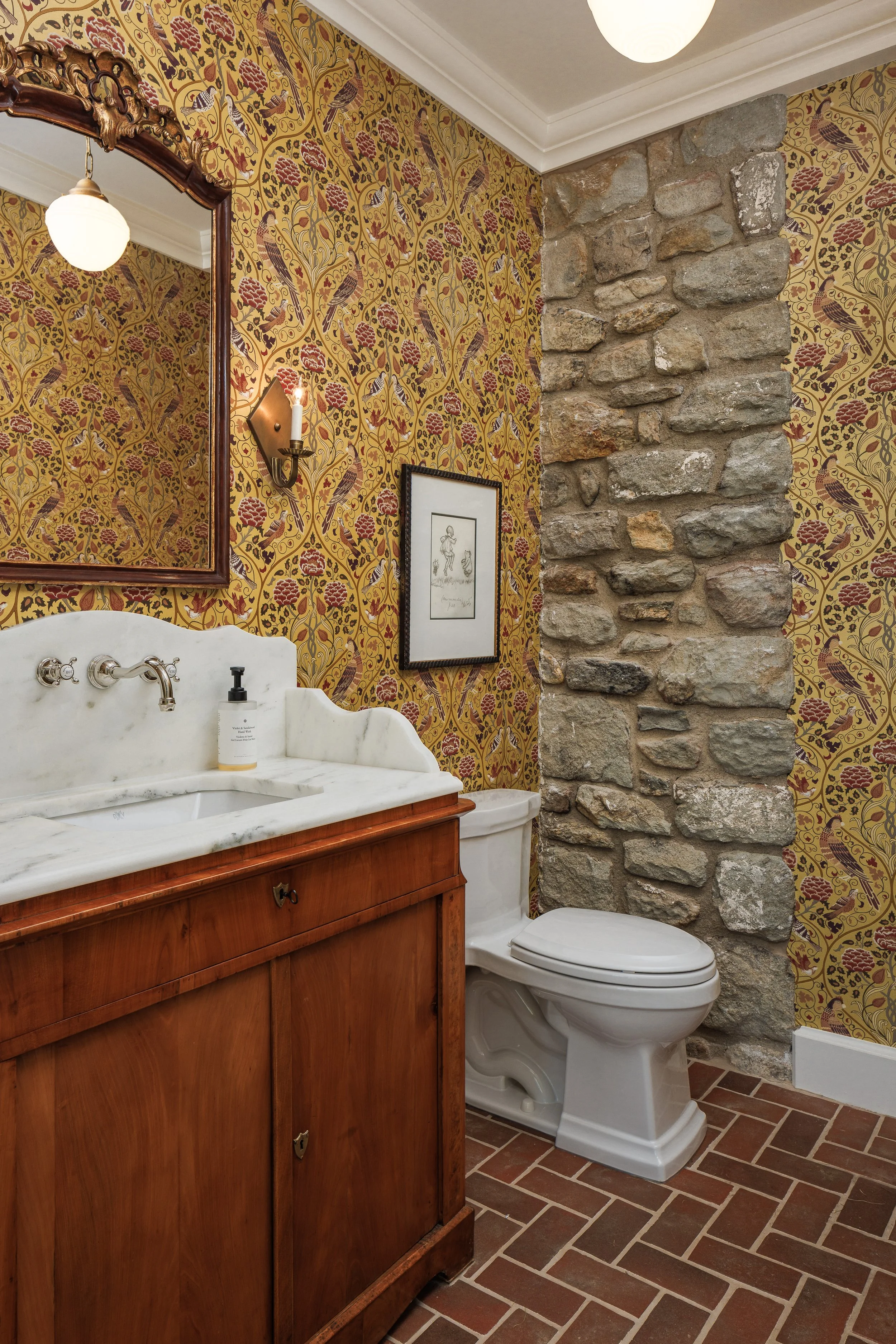 A vintage-style bathroom with a stone wall, a wooden vanity with a marble countertop, a mirror, a framed picture, and a toilet, featuring warm yellow patterned wallpaper and brick flooring.