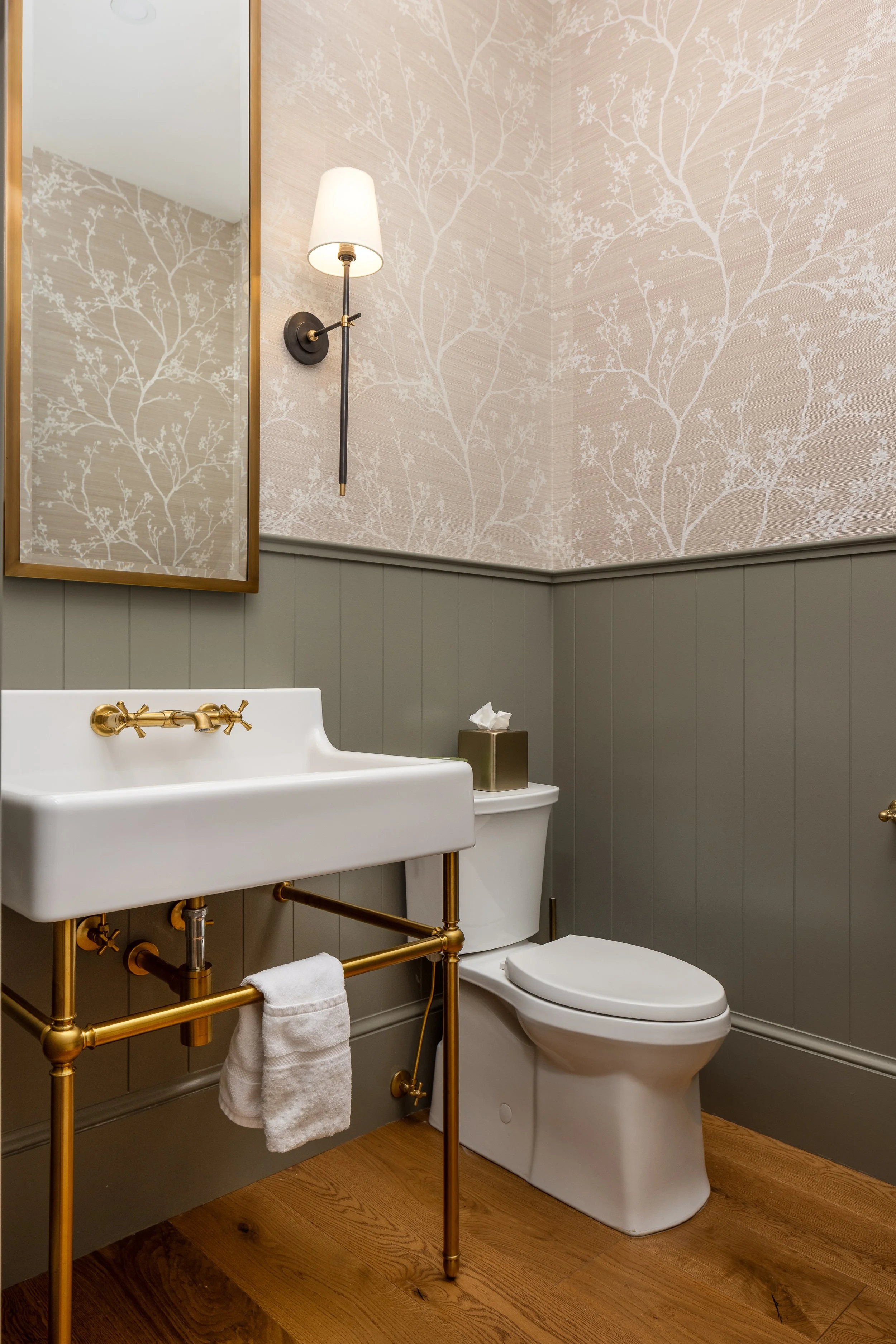 A bathroom with a white sink and gold fixtures, a toilet, a wall-mounted mirror, a wall light, tissue box, and wood flooring.