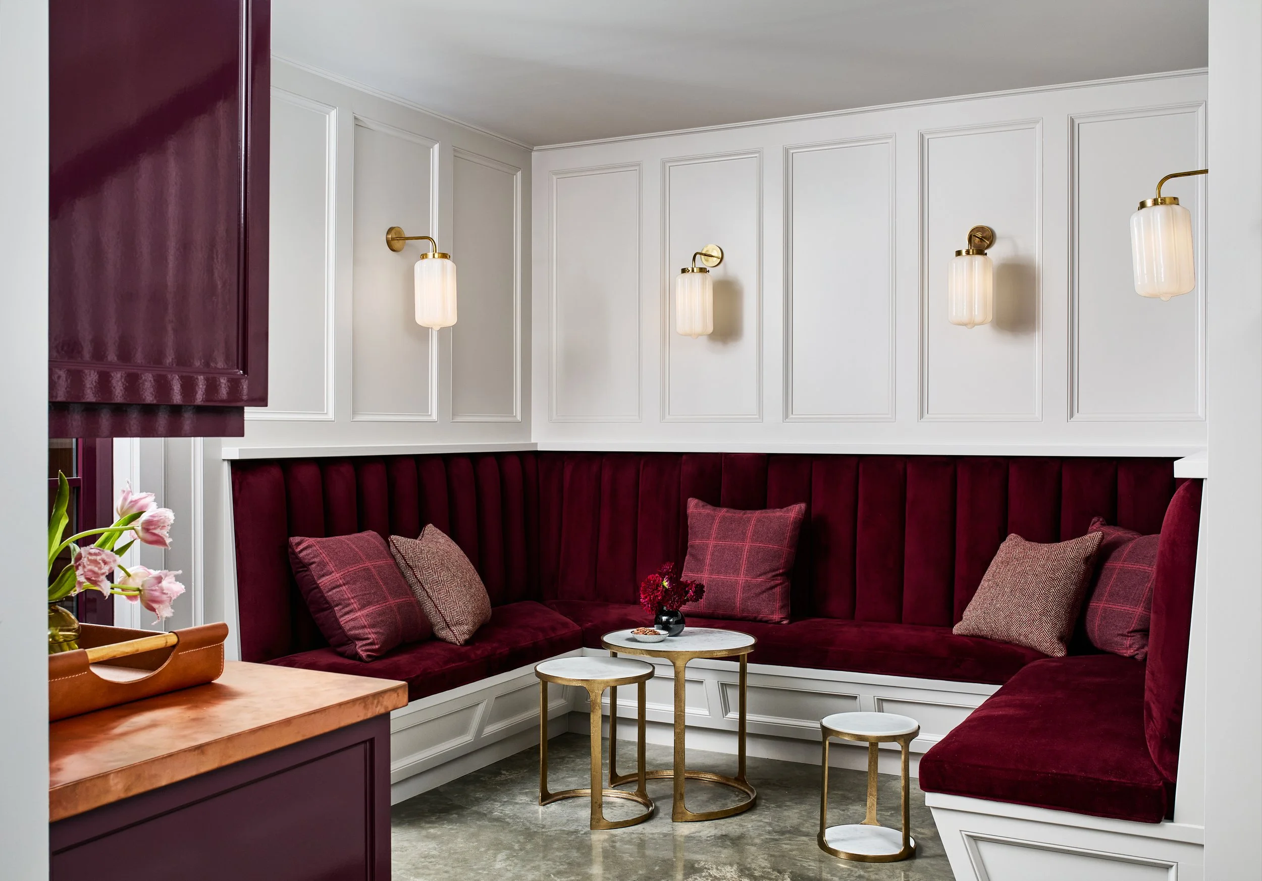 Cozy corner with maroon velvet banquette seating, four decorative pillows, three gold and marble side tables, and three wall sconces with glass shades in a white paneled wall space.