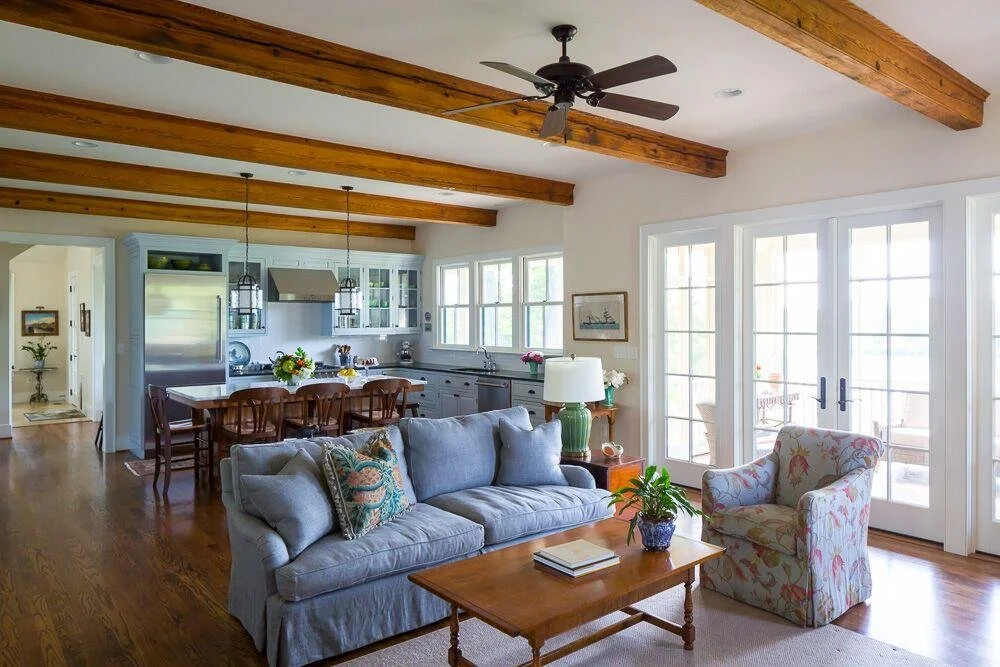 Bright, airy living room with a light blue sofa, floral armchair, wooden coffee table, and large windows with patio doors, open to a kitchen with a dining table and modern appliances, exposed wooden ceiling beams, and hardwood flooring.