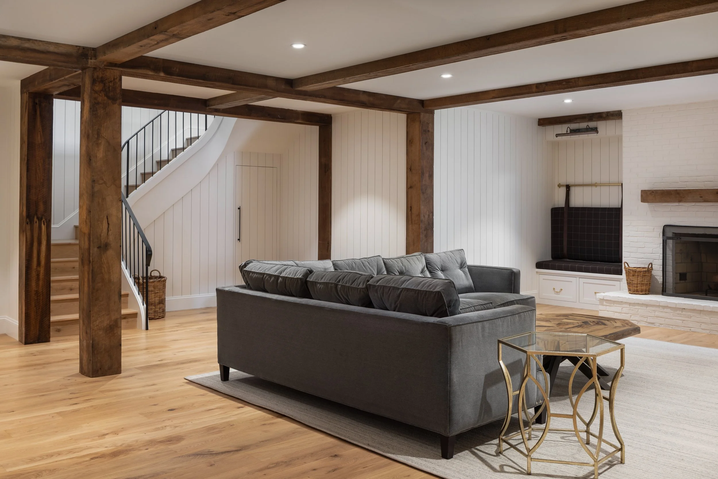 Living room with a gray sofa, a glass side table, a fireplace, wood beams on the ceiling, and a staircase with a curved railing.