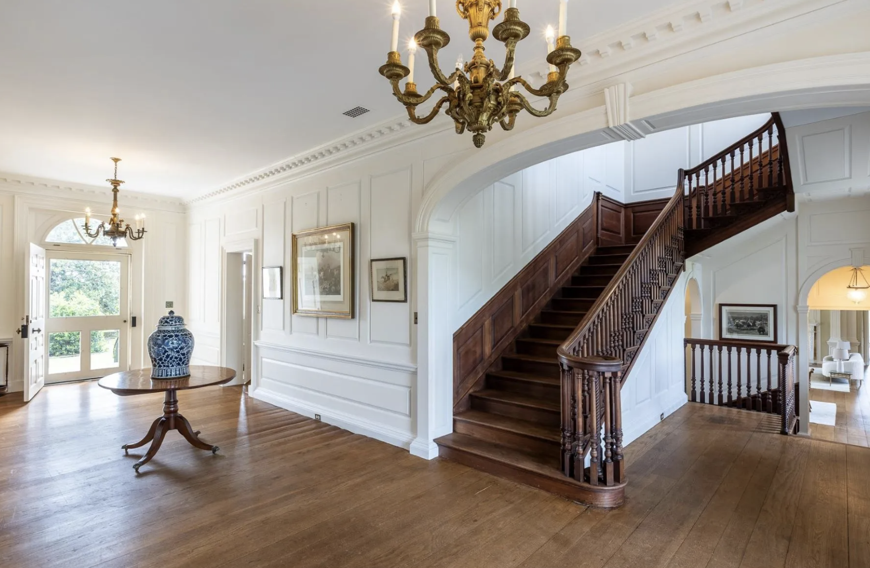 Interior of a grand house with wooden staircase, chandelier, wall art, and wooden flooring.