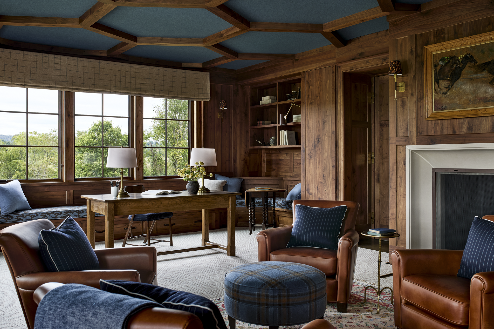 A cozy living room with wood-paneled walls, large windows showing trees outside, leather armchairs with pillows, a wooden table, bookshelves, and wall lamps.