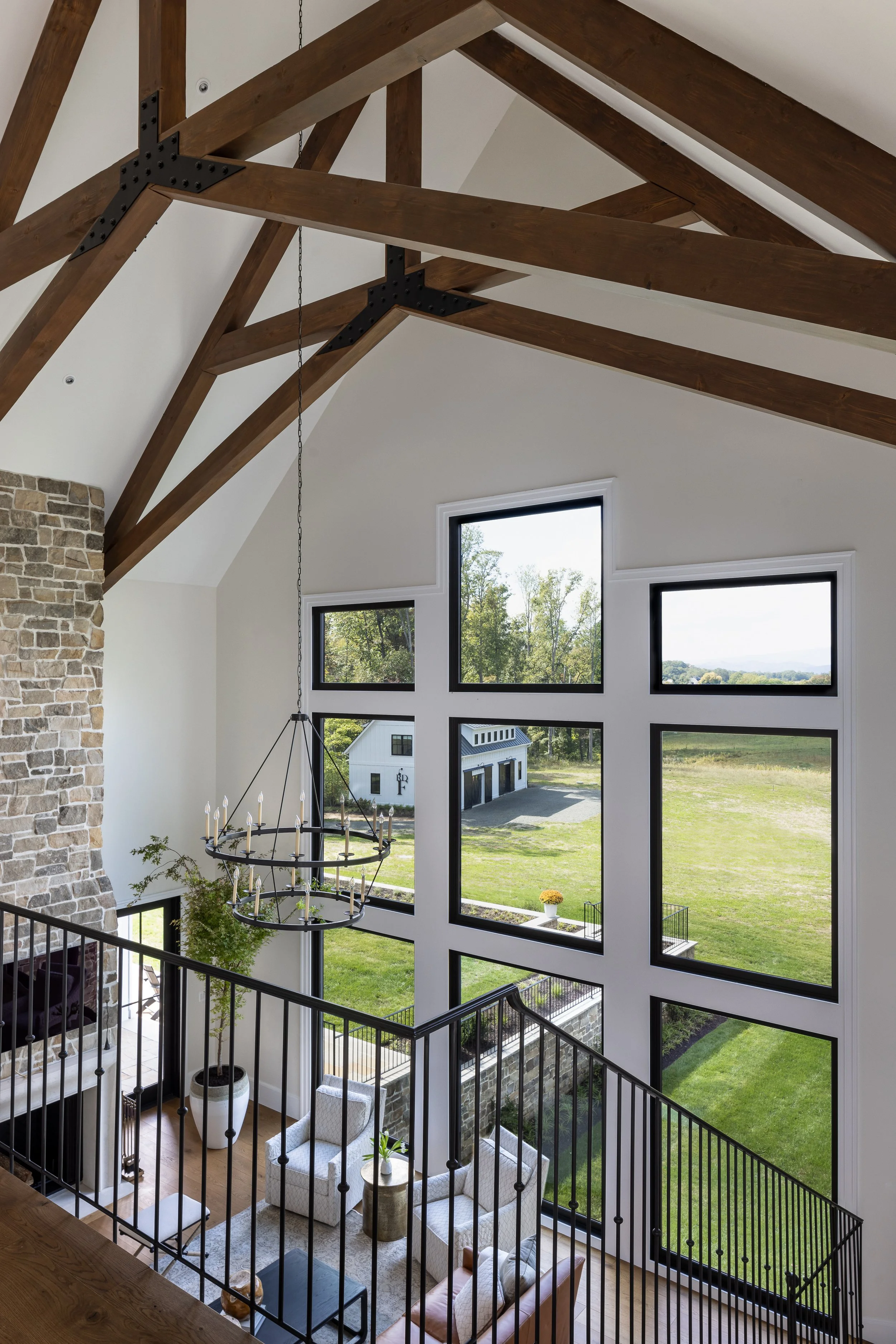 Interior view of a living room with high ceiling, large windows, a stone fireplace, and wooden beams, overlooking a green yard.