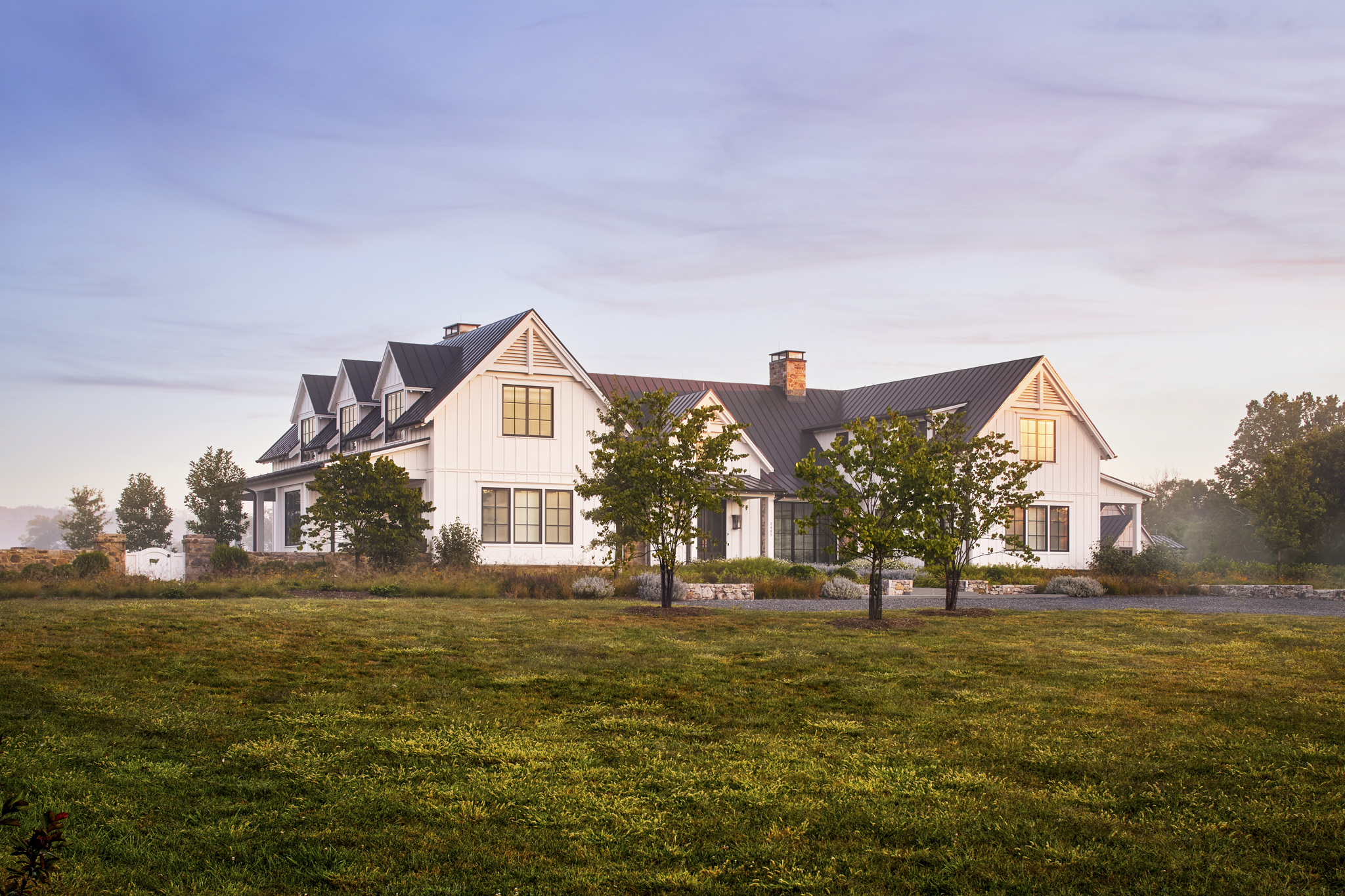 A large white house with black roof and multiple gables, surrounded by trees and grass at sunrise or sunset.
