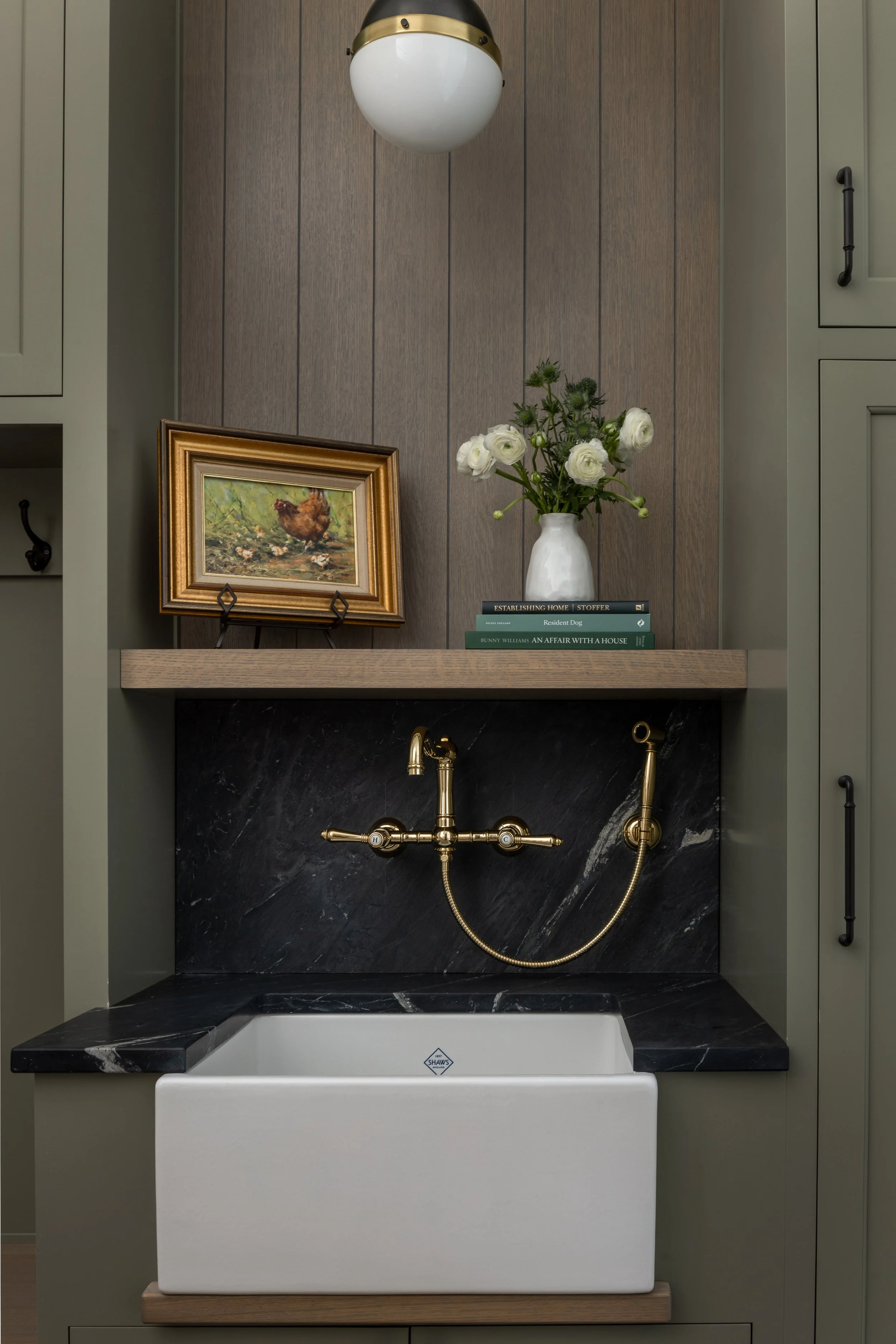 A laundry room sink with black marble countertop, gold faucet, and a farmhouse style white basin, above which there is a wooden shelf with a painting, a vase of white flowers, and a few books. The background features wood and green cabinetry and a gl