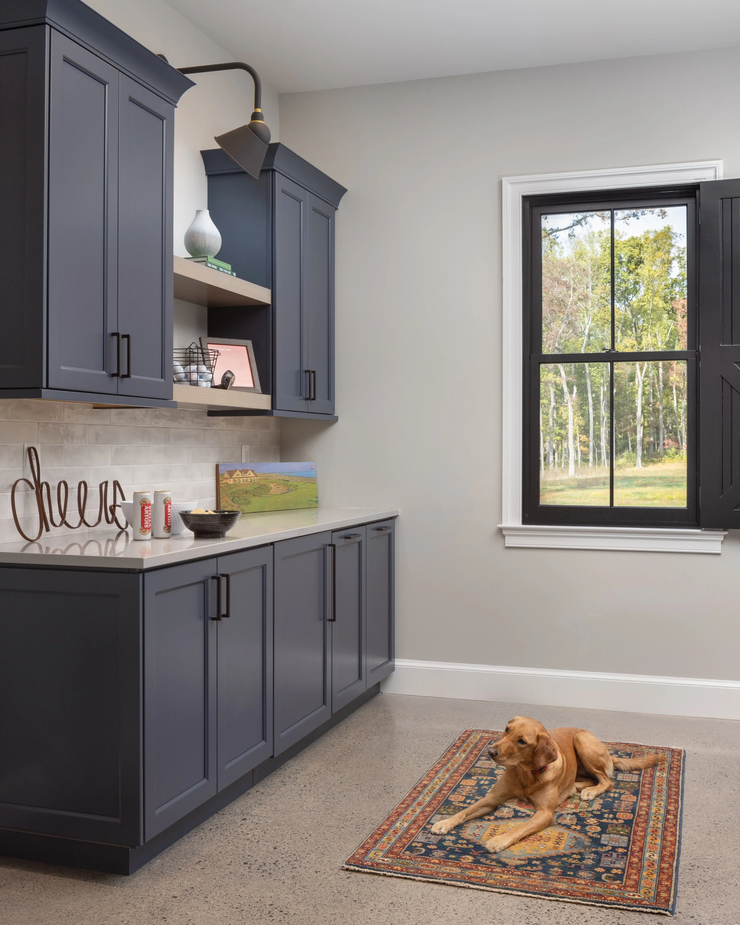 A kitchen with navy blue cabinets, white walls, a window showing a green outdoor landscape, and a dog lying on a patterned rug on the floor.