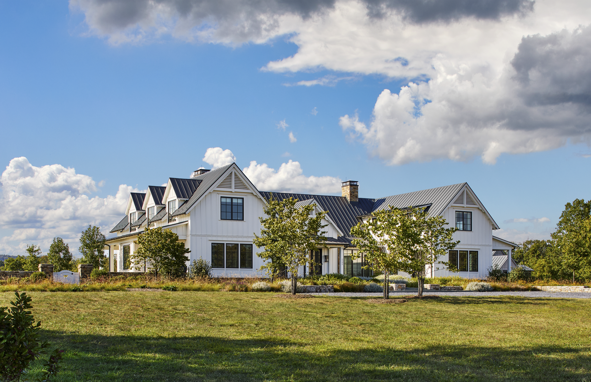 Large white house with black roof and multiple gables, surrounded by trees and a landscaped yard, under a partly cloudy sky.