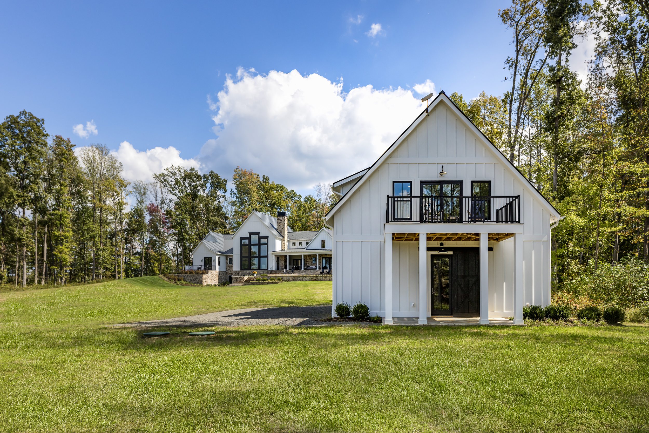Large white barn-style house with black accents on the door and railing, situated on green grass with a wooded area in the background and partly cloudy sky.
