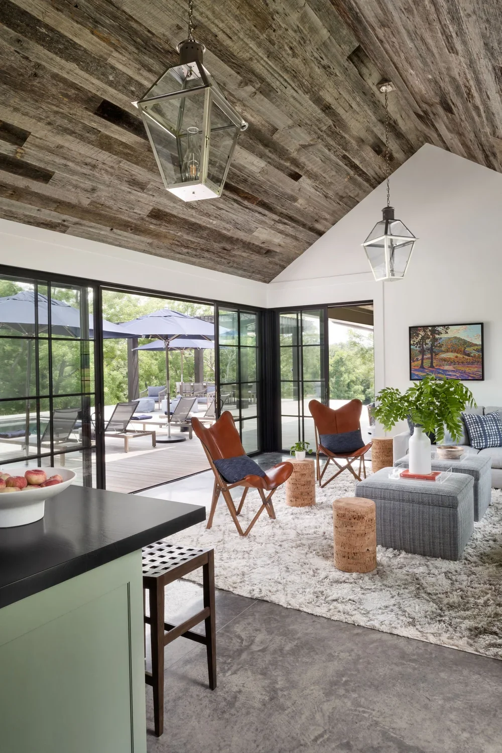 Living room with two hanging glass lantern-style chandeliers, a white sofa with blue pillows, two brown leather chairs, cork ottomans, a gray modern ottoman, a green plant in a white vase, a framed landscape painting, large glass sliding doors leadin