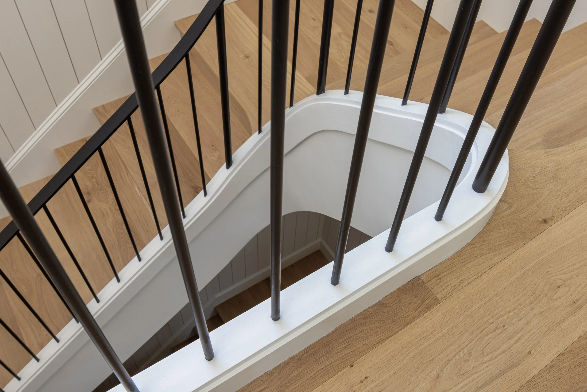 View of a staircase from above showing black railings, wooden steps, and a white curved wall at the bottom with a wooden floor.