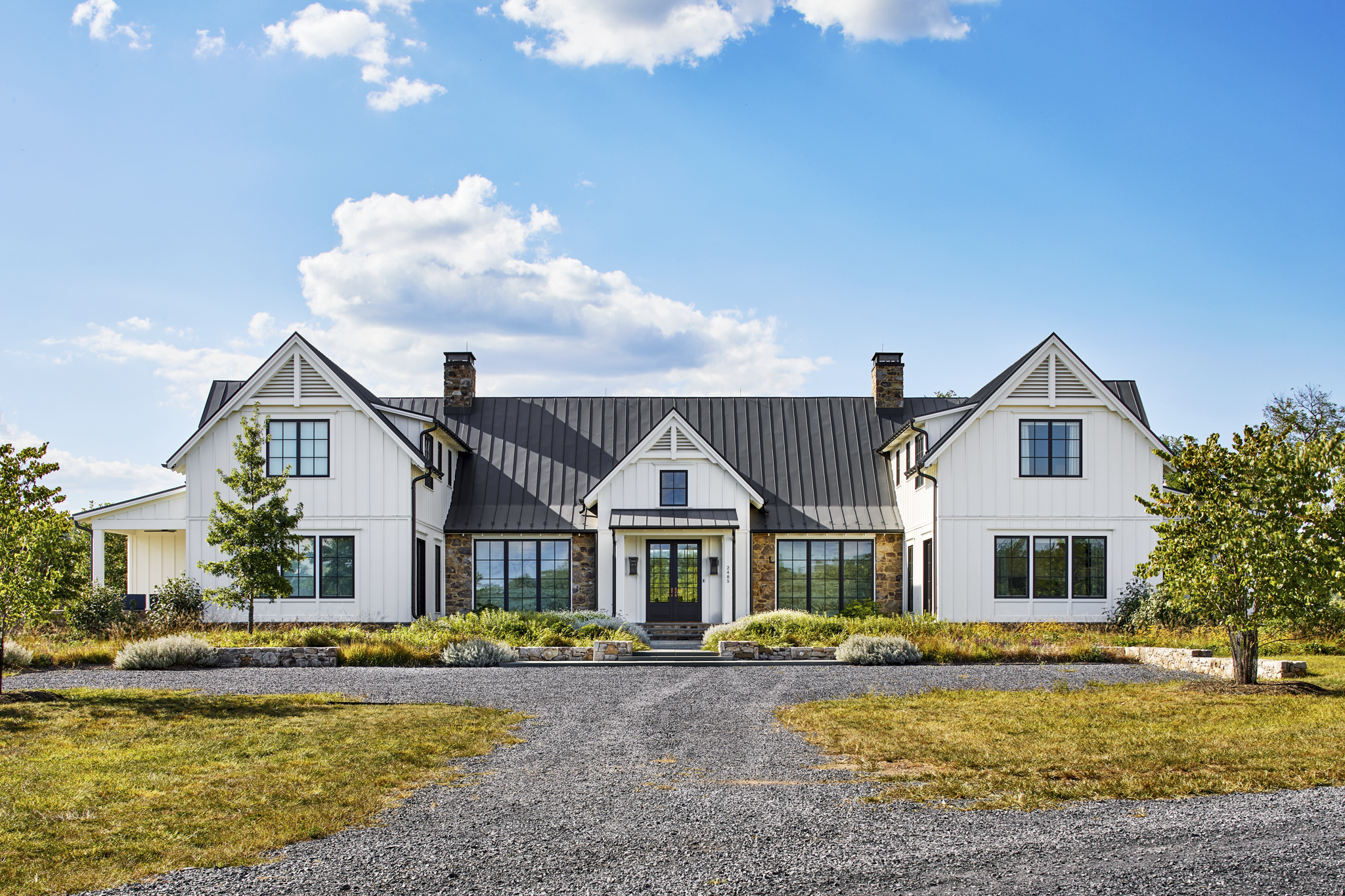A large white house with a black roof, multiple chimneys, and large windows, surrounded by a landscaped yard with trees and shrubs under a partly cloudy sky.