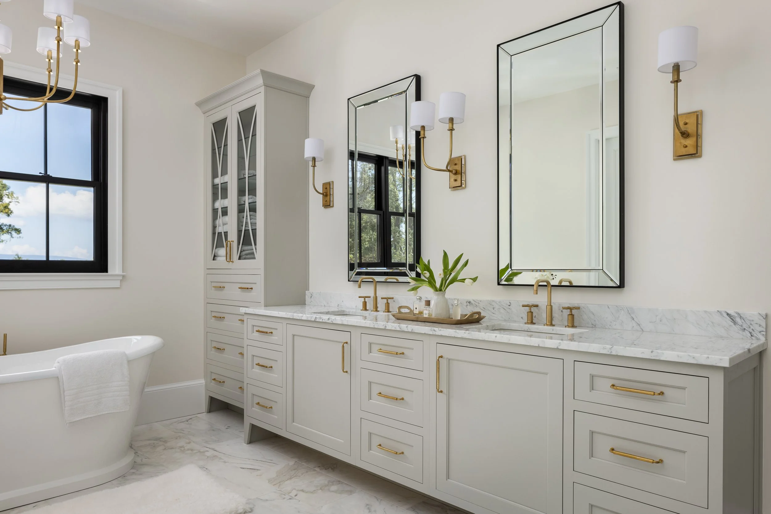 A luxurious bathroom featuring a white double vanity with a marble countertop, gold faucets, and matching gold hardware on white cabinets. Two large mirrors with black frames are mounted above the sinks, flanked by modern wall sconces. A white freest