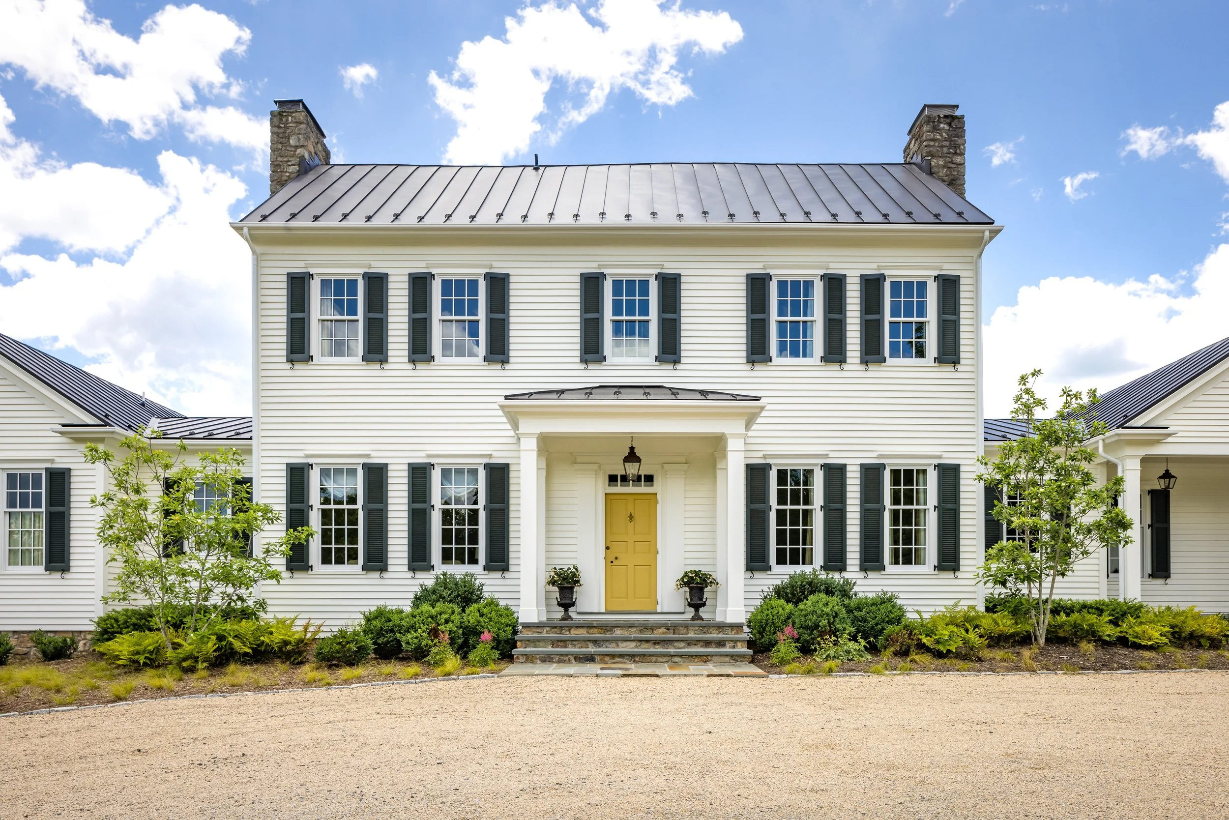 Front view of a two-story white house with black window shutters, a yellow front door, and landscaping in the front yard, under a cloudy sky.